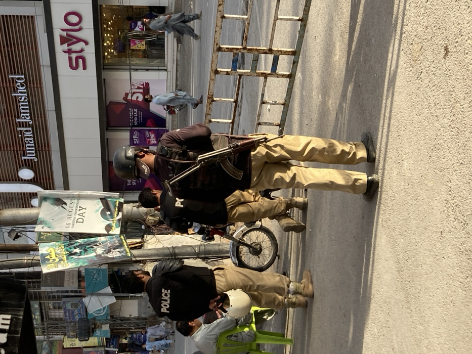 This image depicts an urban street scene during daylight in the city of Milozo, Pakistan. In the foreground, three male individuals in black "POLICE" shirts and khaki trousers are present. Two officers are facing away from the camera; the one on the far right wears a black helmet with "EMC" and "POLICE" markings, and has an AK-style rifle with a wooden stock and magazine slung across his back. Metal barriers define the immediate foreground on the right. To the left of these two officers, a third officer is partially visible, holding a white motorcycle helmet in his left hand. A black motorcycle is parked near this officer. Further left, a man in a light-colored shirt is seated on a bright green plastic chair.
In the mid-ground and background, various elements of a bustling commercial street are visible. A utility pole on the left is adorned with multiple cables and several banners, including one clearly stating "14 AUGUST PAKISTAN INDEPENDENCE DAY" with a Pakistani flag design, and another with Urdu text. Across the street, two prominent commercial establishments, "Junaid Jamshed" and "Stylo," are visible with their signage. The "Stylo" store displays sale advertisements, including "THE GREAT stylo SALE 51%". Several pedestrians, mostly men in traditional attire, are walking on the sidewalk and crossing the street in the background. The scene is illuminated by natural light, casting shadows from the subjects onto the light-colored paved ground.