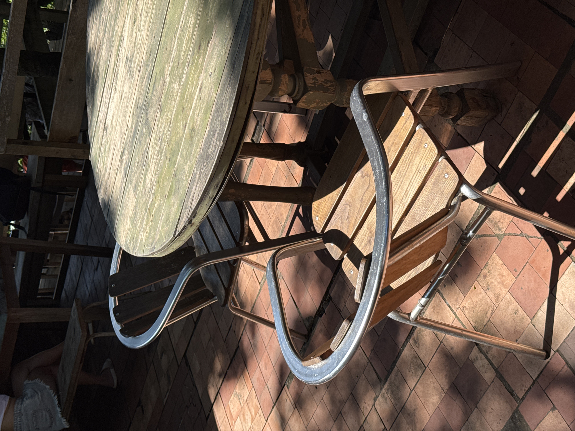 This is a photograph of an outdoor seating area in Buknari, Georgia. Two wooden chairs with metal armrests are positioned in front of a weathered wooden table. The table has a round, planked surface with a greenish hue and is supported by ornate wooden legs. The chairs are placed on a tiled patio, which is partially illuminated by sunlight, creating dappled shadows. In the background, a person is seated on a wooden bench, their legs crossed, wearing a denim skirt and white shoes. The overall scene suggests a casual outdoor dining or lounging space.