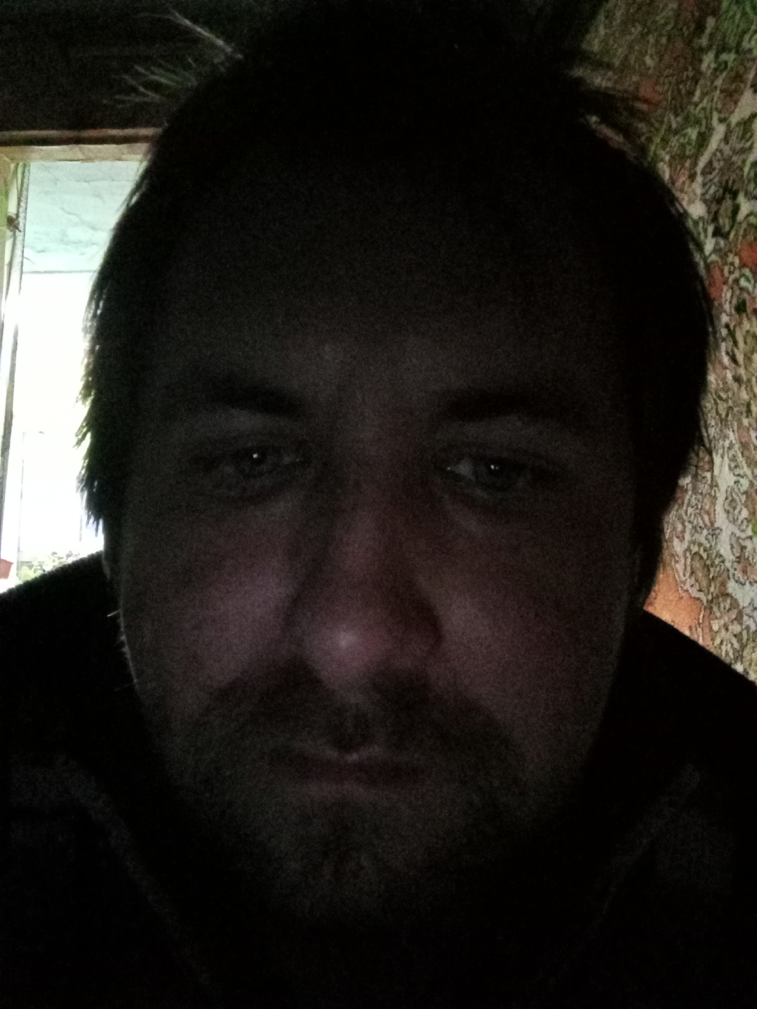 A close-up, dimly lit photograph shows a man with dark hair and a beard looking down. He appears to be indoors; a window is visible in the upper left, and a patterned wall covering is seen on the right. There's no explicit information linking this image to the city of Parabel, Russia.