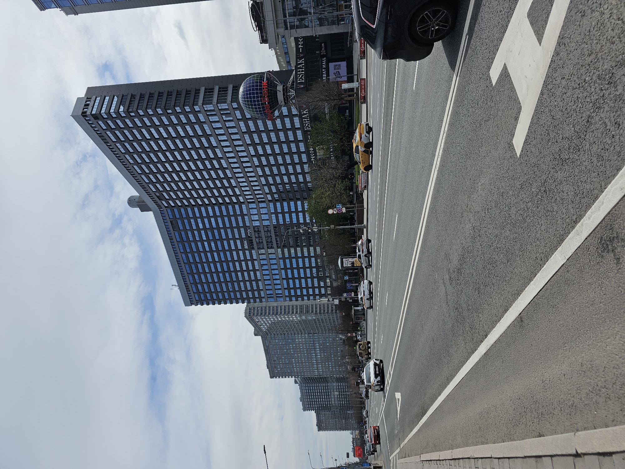 The image shows a street in Moscow, Russia, lined with tall buildings and a busy roadway.
The buildings are modern, high-rise structures with grid-like window patterns. A prominent building on the left features a large globe-shaped structure near its top. A sign on the building reads "ESHAK" and "ARBAT HALL CLUB."
The roadway is wide, with multiple lanes of traffic moving in both directions. Vehicles, including cars, taxis, and trucks, are visible. The road markings are clear, including lane dividers and directional arrows.
The sky is overcast, with clouds visible in the background.