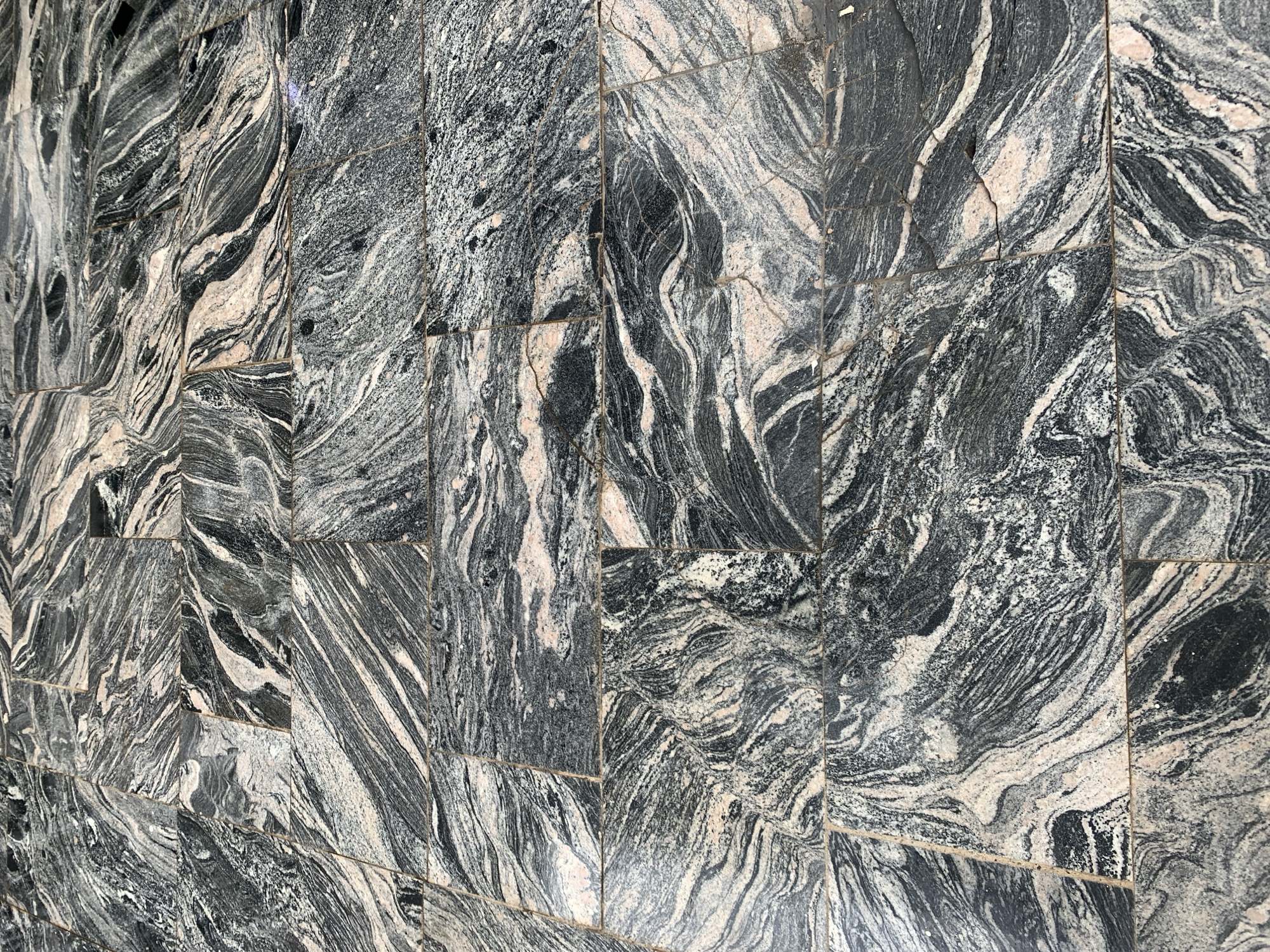 The image displays a close-up, overhead view of a stone-tiled floor. The tiles are rectangular and laid in a grid pattern. The stone itself is a dark grey with prominent white and pinkish veins, giving it a marbled or granite-like appearance. The pattern of the veins is organic and swirling, with some areas exhibiting finer lines and others broader, more fluid shapes. The grout lines between the tiles are visible and appear to be a light grey color. There are some minor cracks visible in some of the tiles, particularly noticeable in the center and right portions of the image. The lighting suggests an outdoor environment, possibly under natural daylight, as there are subtle shadows and variations in brightness across the surface. The scene is static and focuses solely on the texture and pattern of the floor. There are no people, animals, or other objects visible in the frame, nor any indication of the specific location within Gura Top, Nigeria, beyond the fact that it is a tiled surface.