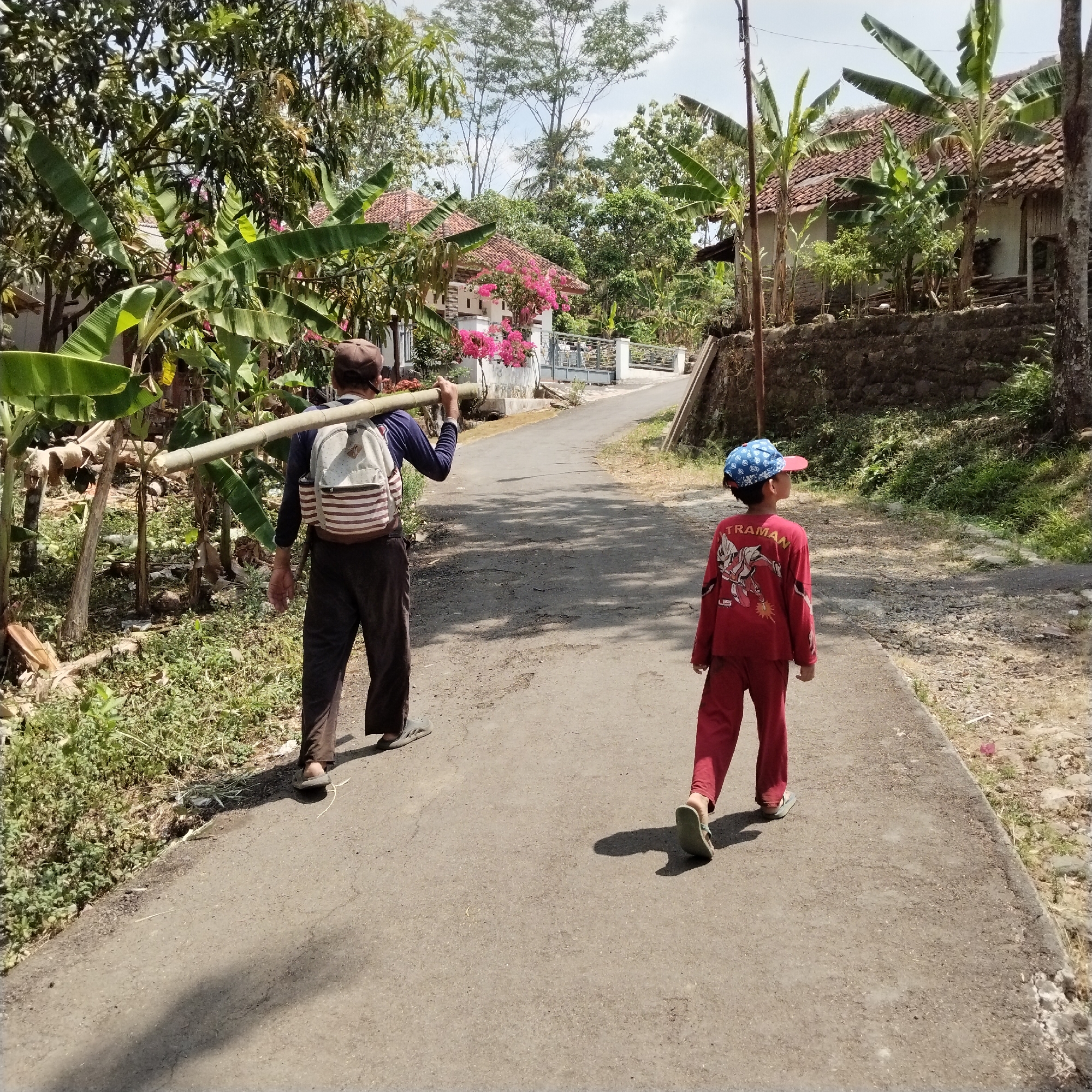 A man and a child walk down a paved road in a rural area. The man, from the back, carries a long bamboo pole across his shoulders. He wears a dark long-sleeved shirt, dark pants, a brown cap, and sandals. A backpack with red and white stripes is on his back. The child, also seen from the back, wears a red long-sleeved shirt with a graphic on it, red pants, and sandals. He has a blue and red patterned baseball cap on. Banana trees and houses with red tile roofs are visible on either side of the road. The scene is lit by sunlight, casting shadows on the road. The location is the city of Karangbaru, Indonesia.
