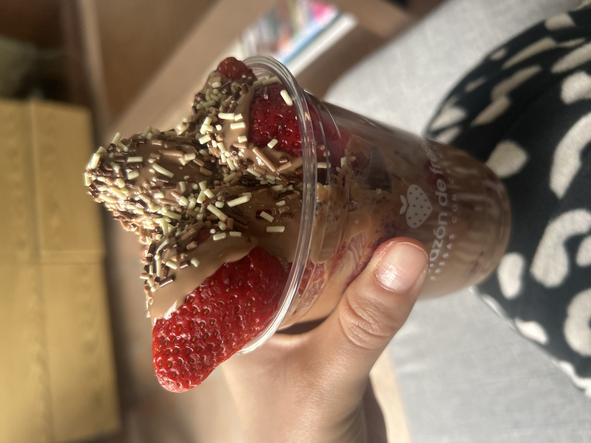 A photograph depicts a clear plastic cup filled with strawberries and chocolate, held in a person's hand. The strawberries appear to be dipped in a light-brown chocolate sauce and topped with sprinkles. The cup features the words "Corazon de fresas" and an image of a strawberry with a heart-shaped center. The person holding the cup has light skin and neatly trimmed fingernails. The background includes a tan surface and a black-and-white patterned surface. The image suggests the location could be a cafe or home setting where the dessert is enjoyed. The city and specific location remain unknown.