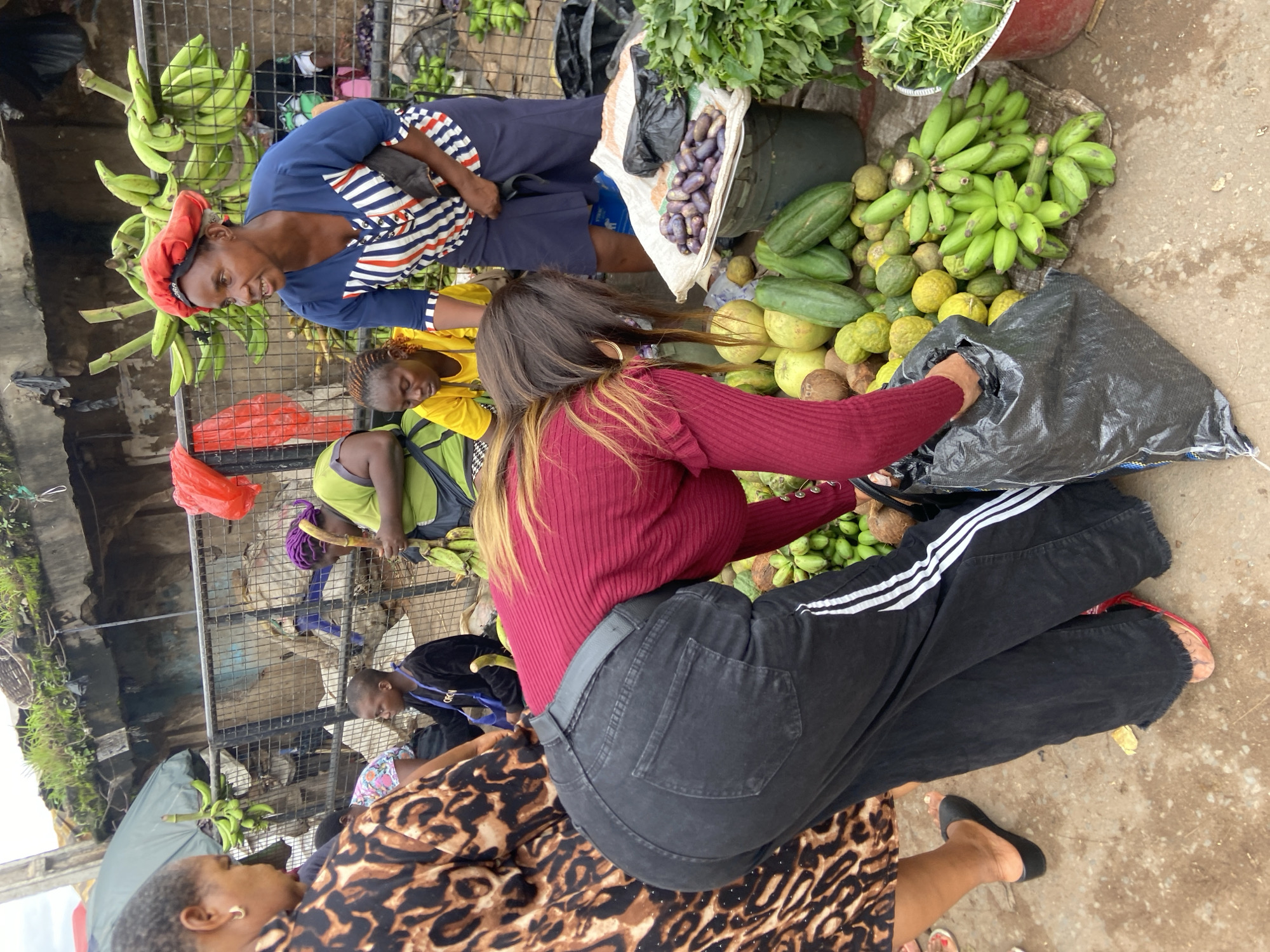 This image captures a vibrant scene at a market stall in Nkpor, Nigeria, likely during daytime given the ambient lighting. In the foreground, a woman wearing a dark red, long-sleeved shirt and black pants is bent over, placing items into a large black plastic bag. Her black jeans have a white stripe detail down the side of each leg and appear frayed at the hem. She wears gold hoop earrings and her long, dark hair is visible. To her left, another woman in a brown and black leopard-print dress is also partially visible, wearing black slip-on shoes.
Behind them, a woman in a blue shirt with white and red stripes and a red headscarf is looking to her right. She is wearing a dark blue skirt. Beside her, a woman in a bright yellow t-shirt is seated, also looking down. A person in a green shirt with purple headscarf is also visible in the background, holding a bunch of green bananas.
The market stall is laden with fresh produce. Prominently displayed are several bunches of green bananas, both hanging and in piles. Other fruits and vegetables include what appear to be gourds, small green fruits, and some dark purple, oval-shaped items in a bucket. A bundle of leafy green vegetables is also visible in a red bucket on the right. The stall is set up outdoors with a metal fence behind the vendors, and a concrete or dirt ground covers the area. The overall environment suggests a bustling outdoor market with a variety of goods for sale.
