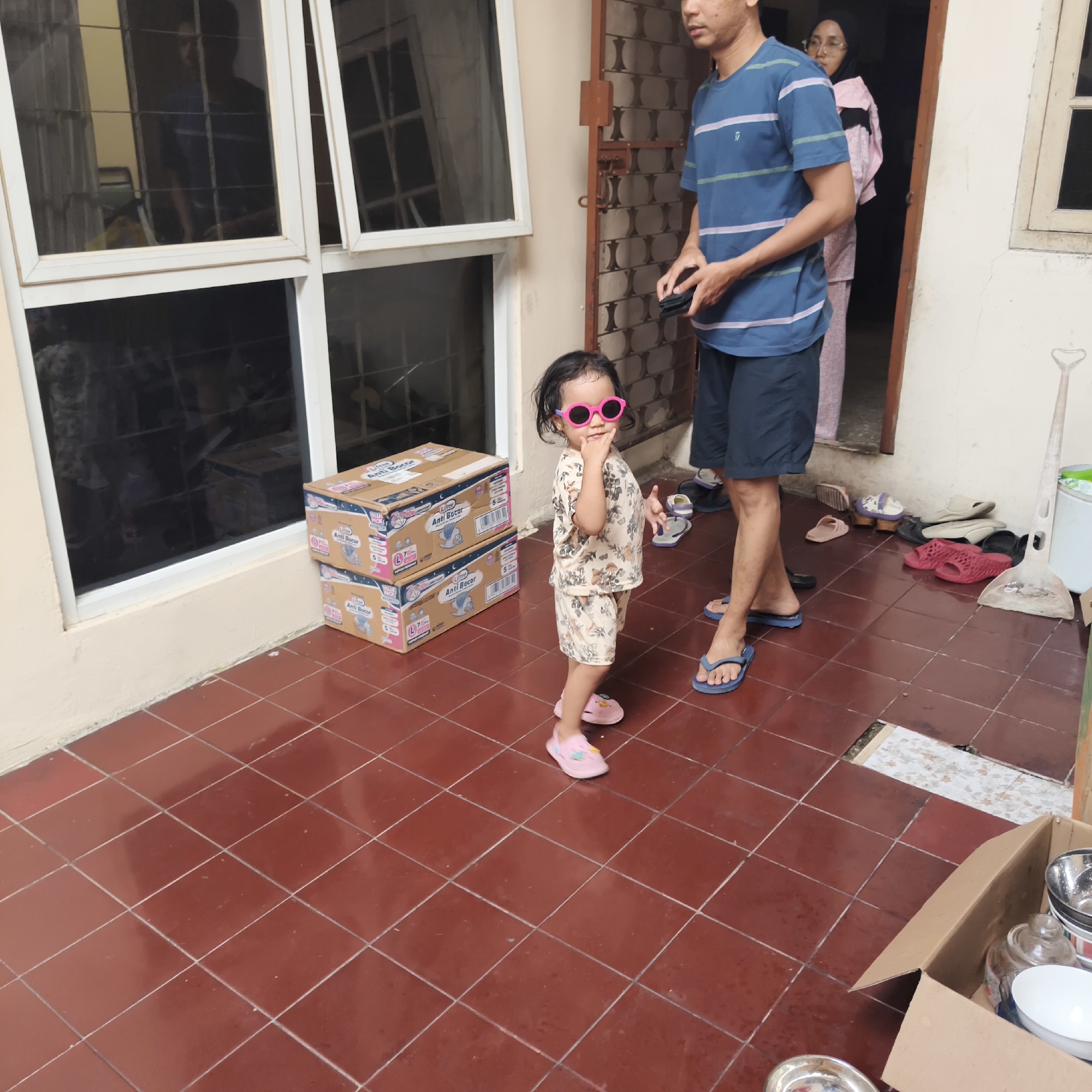 In South Jakarta, Indonesia, a toddler wearing pink sunglasses and a patterned outfit stands on a red-tiled patio, looking towards the camera and touching her mouth. To her left are two stacked cardboard boxes. Behind her, a man in a striped blue t-shirt and dark shorts stands with his hands clasped. Further back, a woman in a headscarf and pink patterned clothing is visible. The patio floor is a grid of red tiles. Various pairs of sandals are scattered on the ground near a doorway and a large dustpan. A partially open cardboard box containing bowls and a glass jar is in the lower right corner. The background includes a wall with a textured pattern, a window with a grid, and a wooden door frame.