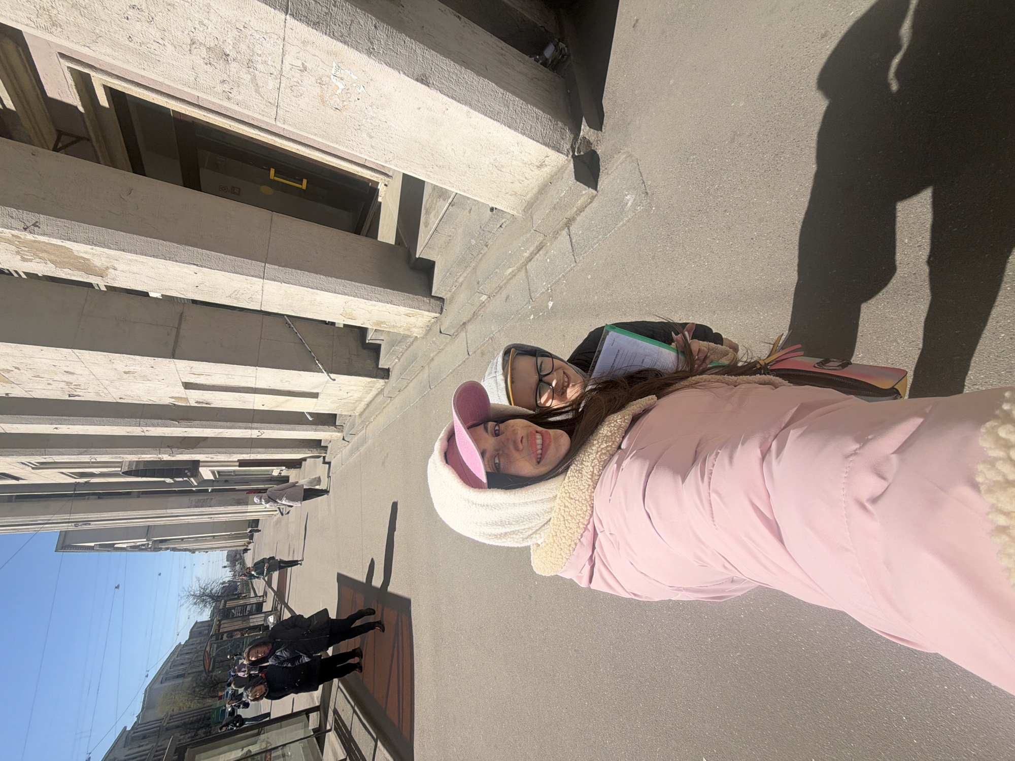 This image captures two young women posing cheerfully on a sunny city street in Saint Petersburg, Russia. The photo, taken at close range, appears to be a casual outdoor snapshot or selfie.
In the foreground, the woman closer to the camera wears a pink puffy jacket with a cream-colored sherpa-lined hood and a pink baseball cap. She is smiling broadly, revealing her teeth. Behind her, another young woman with glasses and a grey hooded sweatshirt is also smiling, holding a green folder or notebook with papers. Both subjects convey a sense of happiness and ease.
The setting is a classic European city street, consistent with Saint Petersburg's architecture. Tall, stately buildings with architectural details like columns line one side of the paved street. The ground is a light grey asphalt or concrete. The sky is clear and bright blue, indicating good weather. Long, distinct shadows are cast on the ground, suggesting it is either morning or late afternoon with bright sunshine.
In the background, other pedestrians can be seen further down the street, and more buildings and trees extend into the distance, contributing to an urban panorama. The overall scene depicts a pleasant day out in the city. No prominent legible text is visible in the image.