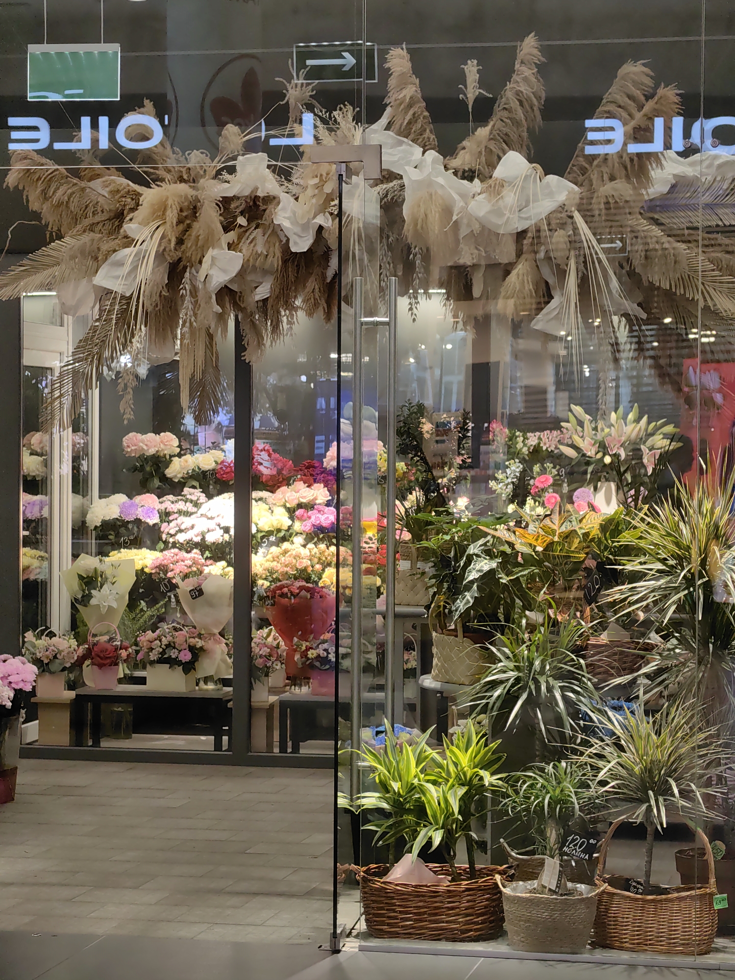 This image depicts the exterior of a flower shop located in Minsk, Belarus. The shop's storefront features a large glass window displaying a variety of floral arrangements and potted plants. Above the display, a decorative arrangement of dried reeds and white fabric hangs from the ceiling. The reflections on the glass show an interior environment with shelves and lights, and exterior signage that includes white text on a dark background.
The window display is richly filled with colorful flowers, including roses, lilies, and gerbera daisies, presented in bouquets and arrangements. Potted plants with green and yellow foliage are also visible in the foreground, housed in woven baskets. The flooring outside the shop is paved with rectangular tiles. The overall impression is of a well-stocked and attractively presented flower shop.