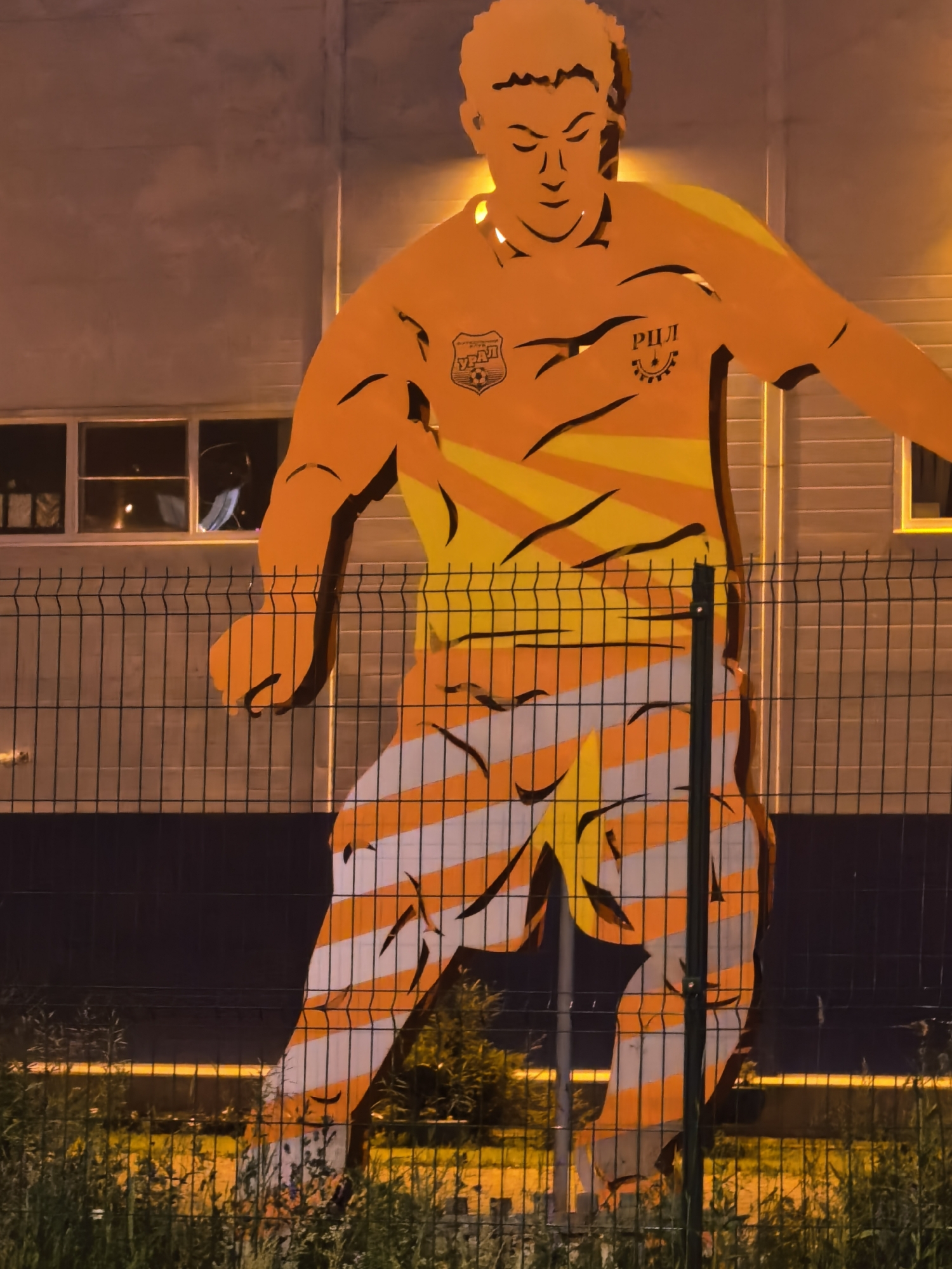 The image displays a large, orange and yellow mural of a soccer player on a building's exterior wall. The mural is illuminated by a warm light, casting a glow on the building's textured facade. A chain-link fence runs across the foreground, partially obscuring the mural. The lower portion of the mural features a pattern of orange and white stripes. The soccer player is depicted in mid-action, with an intense expression. A logo, possibly a crest with a soccer ball, is visible on the player's chest. The surrounding environment appears to be an outdoor urban setting at night, with some vegetation visible below the fence.