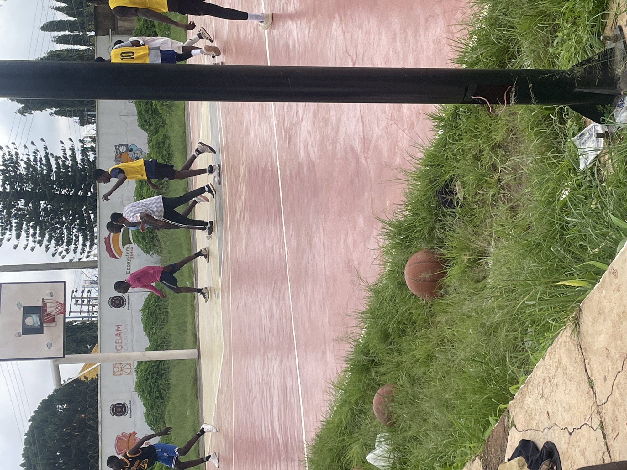 In the city of Jos, Nigeria, a group of young men is playing basketball on an outdoor court. The court is marked with white lines on a reddish-pink surface. A basketball hoop is visible in the background, attached to a white backboard. Several players are wearing yellow bibs over their athletic attire. Some players are in motion, appearing to be engaged in a game. The court is surrounded by a low wall with some faded graffiti. Beyond the court, there is dense green vegetation, including trees and grass. Two basketballs are visible in the overgrown grass near the edge of the court. A dark pole, likely part of the basketball apparatus, stands prominently in the foreground. The sky is overcast, suggesting a cloudy day.