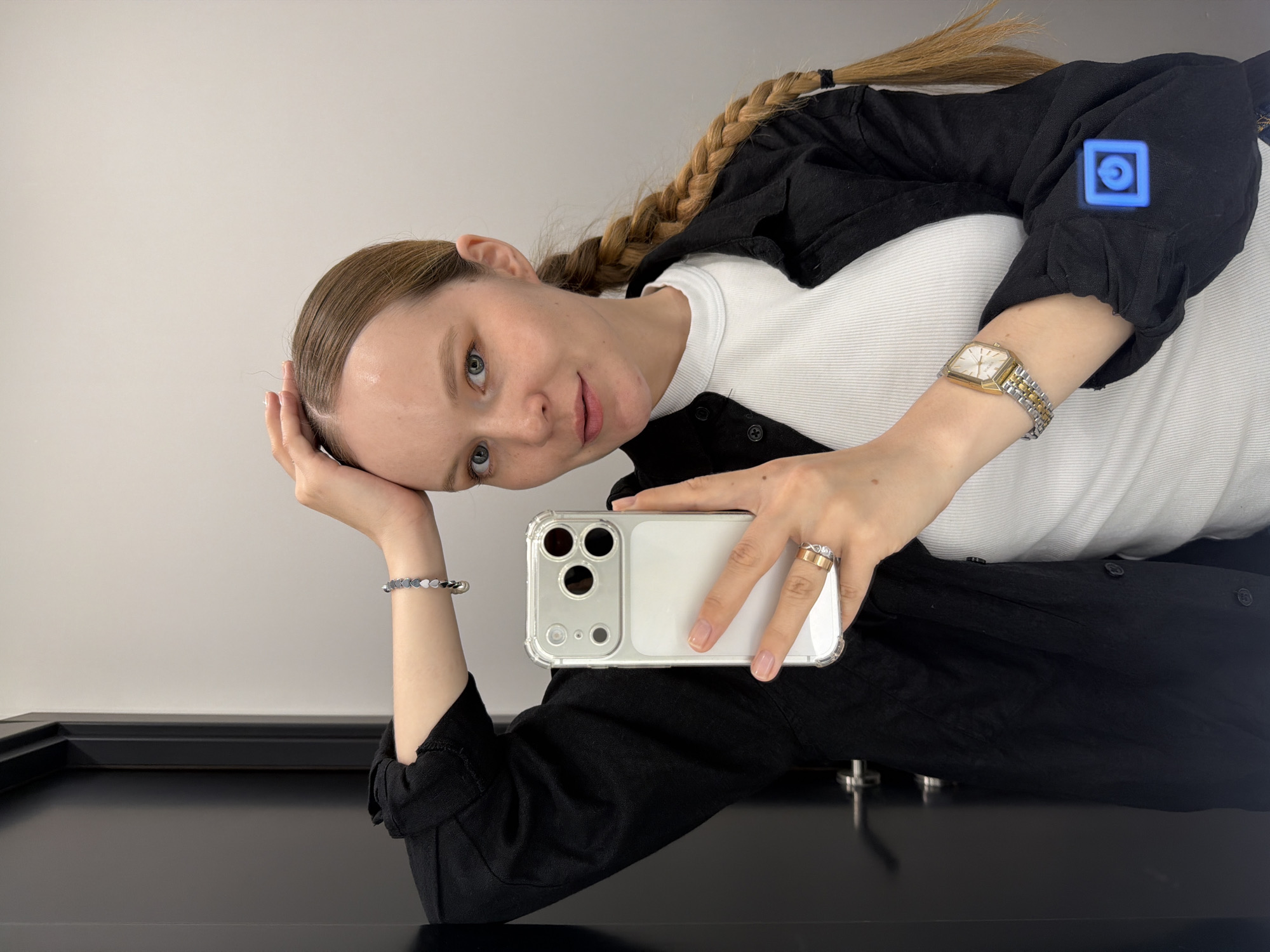A young woman is depicted in a mirror selfie, taken in Izhevsk, Russia. Her head is tilted to her left, resting on her left hand, and she gazes directly at the camera with a subtle, gentle smile. She has light brown hair styled in a long side braid that extends over her right shoulder.
She is wearing a white ribbed long-sleeved top beneath an unbuttoned black shirt. On her right wrist, she sports a rectangular watch with a gold bezel and a two-tone silver and gold band. Her left wrist features a bracelet composed of small, heart-shaped black and white beads. Her right hand, holding a white smartphone in a clear protective case, displays two rings: a wide gold band and a thinner silver band adorned with a stone. The smartphone appears to be an iPhone Pro model, identifiable by its triple-camera system.
The background is a plain, light-colored wall, while a dark, reflective surface, possibly a table or counter, occupies the lower portion of the frame. A glowing blue square icon containing a circular symbol is visible as a reflection on her black shirt, near her right shoulder. The lighting is bright and even, indicating an indoor setting. No visible text is present in the image.