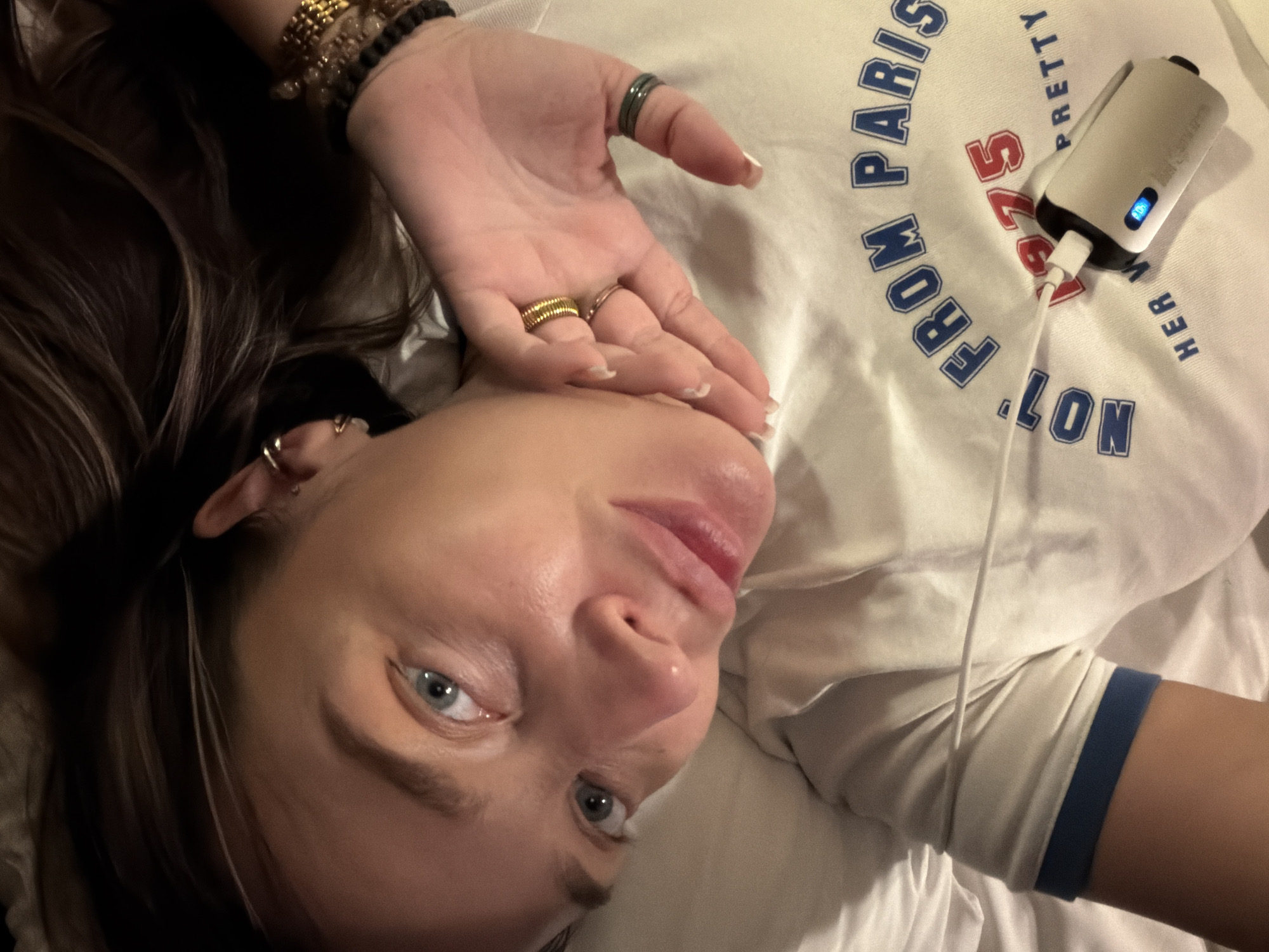 The image shows a woman lying on a white surface, presumably a bed, wearing a white t-shirt with blue lettering that reads "NOT PRETTY HER FROM PARIS 75 LON". She is angled towards the camera with her head resting on the surface. Her right hand is raised near her face. She has several rings on her fingers and bracelets on her wrist. A portable charger is visible on her shirt, plugged in with a white cable. There is no indication of the location being Moscow, Russia.
