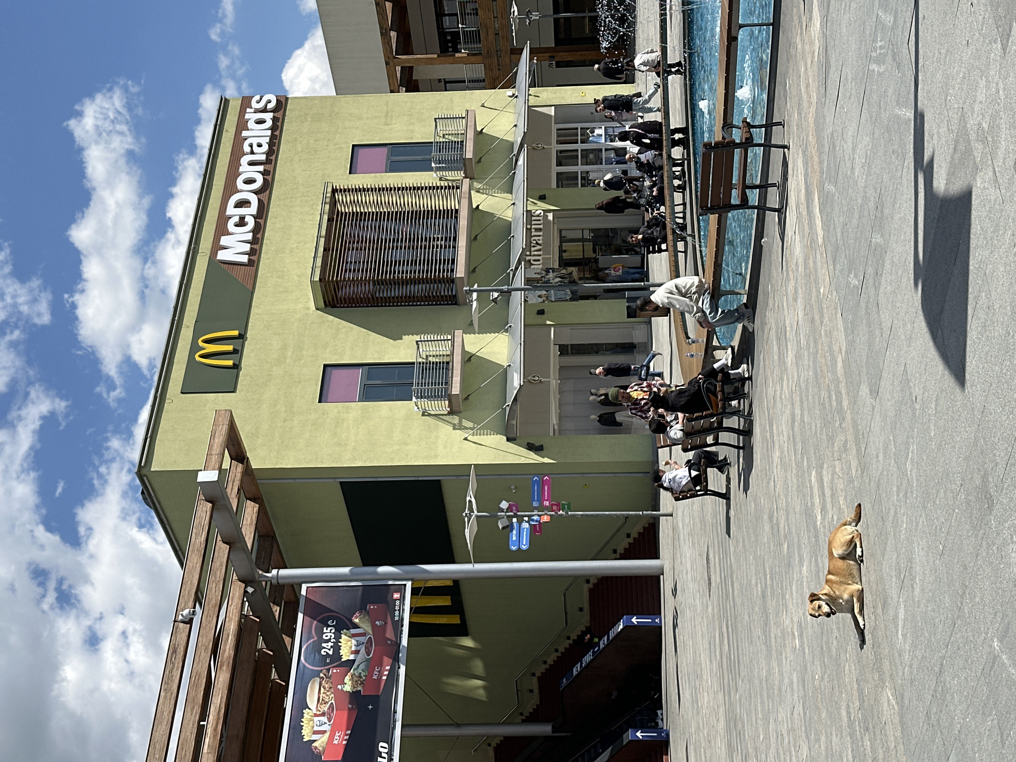 This image depicts a sunny outdoor plaza or commercial area in Tbilisi, Georgia, under a bright blue sky with scattered white clouds. A prominent light olive-green building dominates the scene, featuring a large "McDonald's" sign and its golden arches logo on its upper facade. Below, the name "Stradivarius" is visible on another storefront. The building incorporates modern architectural elements like balconies with slatted wooden screens.
Numerous people are present, seated at outdoor tables in front of the stores, some enjoying meals or drinks. A long, narrow water feature or fountain runs alongside the seating area, with a person bending down near it. In the foreground, a light brown dog is lying relaxed on the light grey paved ground. The strong shadows cast by people and structures indicate a daytime setting, likely midday or early afternoon.
Visible text includes the "McDonald's" and "Stradivarius" signs. A large billboard on the lower left advertises KFC products, showing food items, a price of "24.95 €", and operating hours from "10:00-01:00." Directional signs on a pole point towards "entrance" and "parking." The scene conveys a bustling yet relaxed atmosphere of an urban shopping and dining destination.