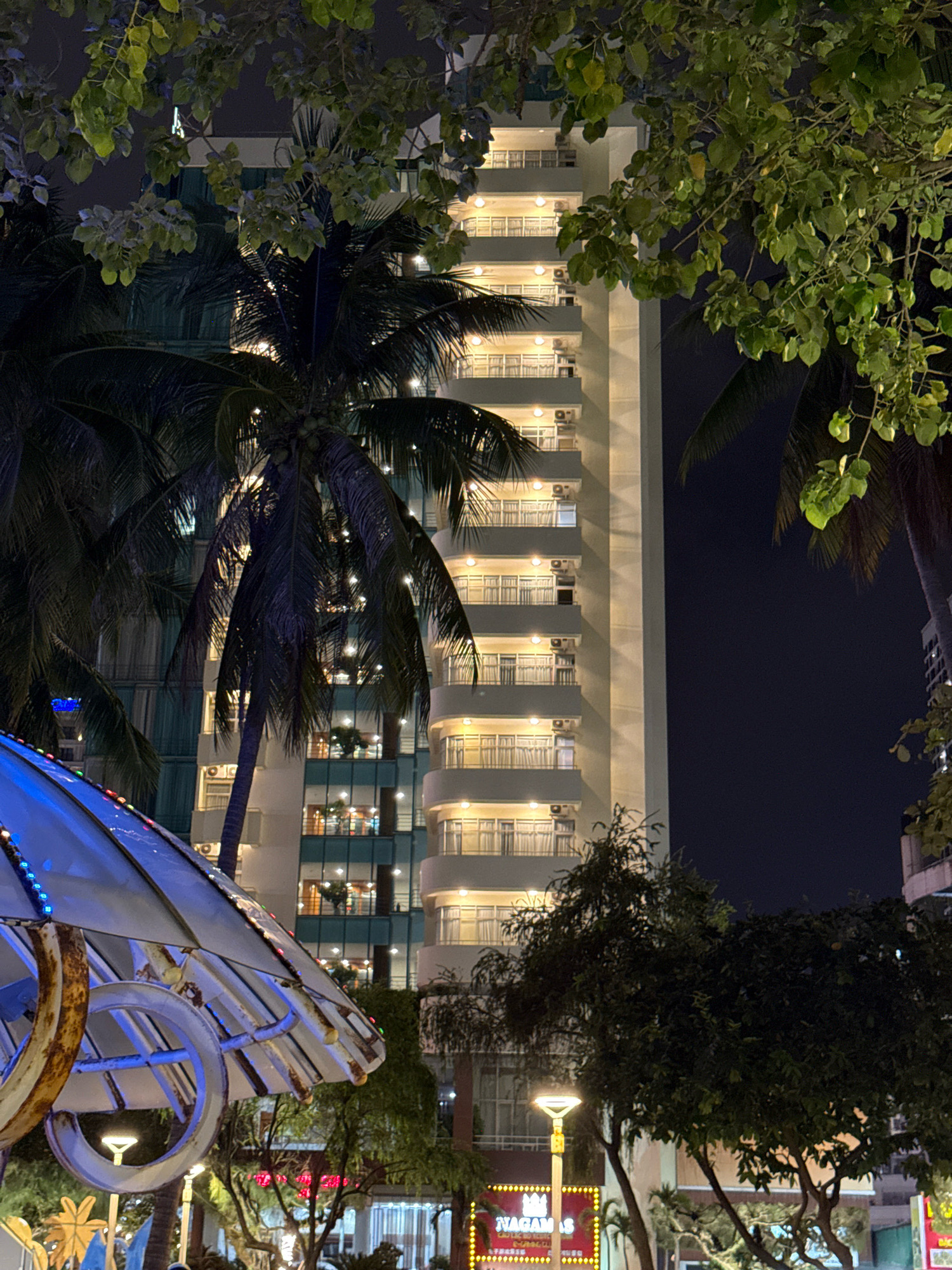 In Nha Trang, Vietnam, at night, a tall, brightly lit building is visible behind palm trees and lush foliage. The building's facade is illuminated with warm yellow lights from the balconies. In the foreground, a portion of a brightly lit, blue-tinted structure, possibly a canopy or awning with decorative lights, is visible. The scene is a street-level view, with streetlights and signs indicating a commercial or entertainment area. The overall atmosphere is lively and urban, with the natural elements of trees contrasting with the modern architecture.