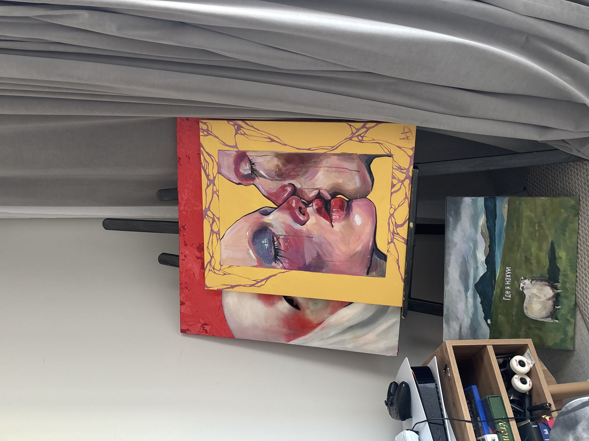 The image shows a cluttered indoor scene under what appears to be a bed. Two paintings lean against the wall: one featuring a stylized depiction of two people kissing with a yellow background and a red edge, and another showing a sheep in a green field with mountains in the background. Russian text is present on the sheep painting. Below these paintings is a wooden organizer containing various small items including what appear to be skateboard wheels, game cases, and other paraphernalia. A PlayStation 5 console and gaming headset are partially visible below the organizer. The photograph was taken indoors, location unknown. There is no evidence to suggest this image was taken in Krasnoyarsk, Russia.
