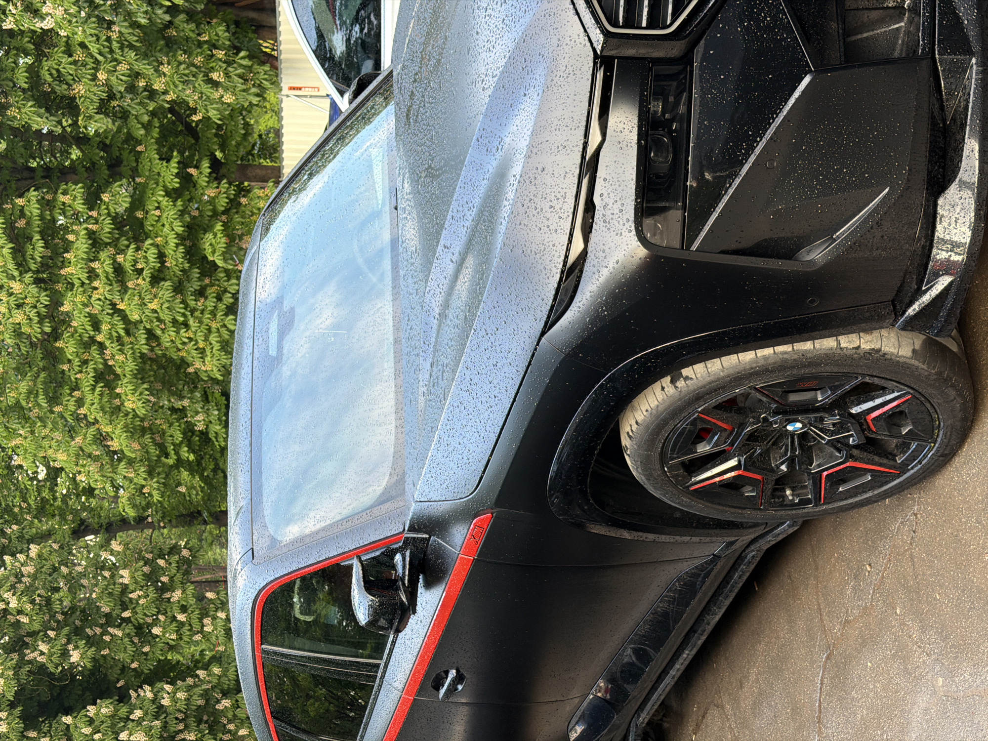 The image shows the front end of a black BMW X6 M Competition car. The car is wet with raindrops, indicating it has been raining. The car has red accents around the windows and on the wheels. The background consists of lush green trees with blooming white flowers. The exact location in Sofia, Bulgaria, cannot be determined from the image.