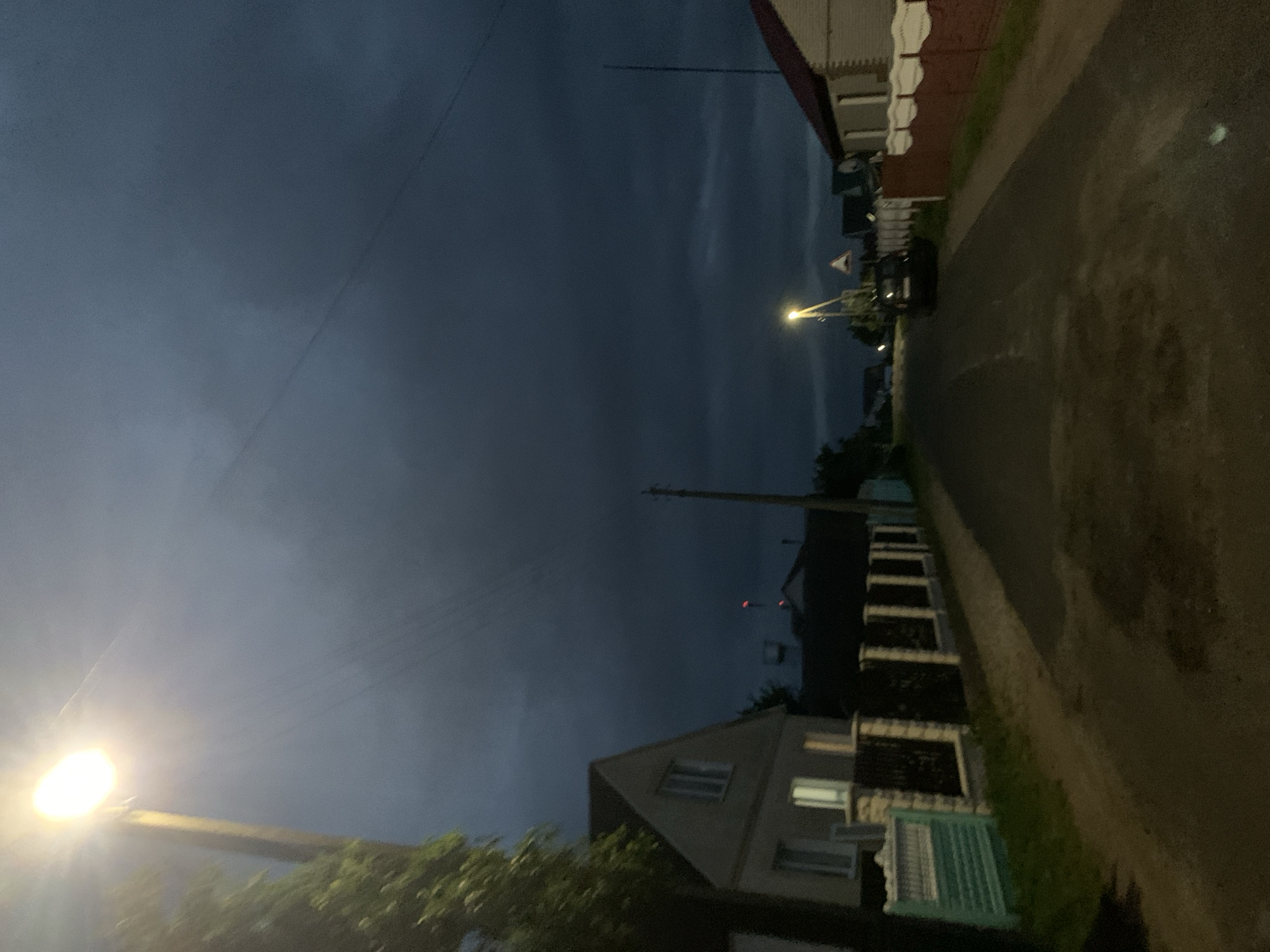 The image depicts a dimly lit residential street at dusk or night, likely in Kapyĺ, Belarus. The sky is a deep, dark blue, with some lighter clouds or atmospheric haze visible. A bright streetlamp illuminates the foreground on the left, casting a strong glow and creating lens flare. A power line stretches across the sky from left to right.
On the right side of the street, a paved road curves away into the distance. Adjacent to the road are several houses with traditional architecture, featuring pale walls and dark roofs. Some of the houses have illuminated windows, suggesting occupancy. A black car is parked on the side of the road. Fencing and greenery are also visible along the street.
The overall mood of the image is quiet and slightly mysterious, due to the low light conditions. The time of day appears to be after sunset, with artificial lighting taking over. No people are clearly visible in the image. There are no visible text elements.