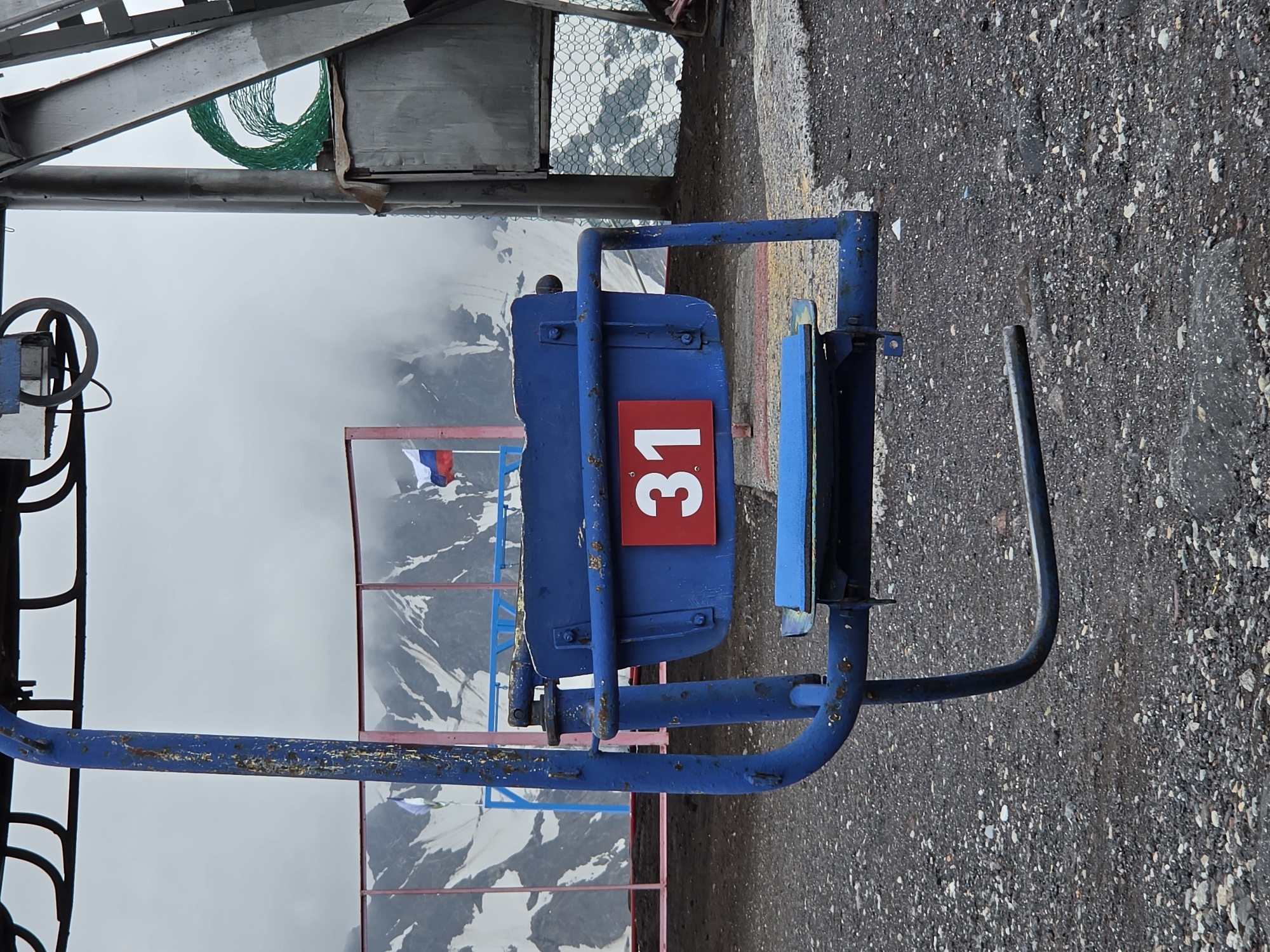 The image captures a ski lift chair in the foreground, painted blue with visible rust and wear. A red rectangular sign with the white number "31" is affixed to the chair. The background shows a cloudy sky over snow-capped mountains. To the left, a portion of a fence with a pink railing is visible, with Russian flags flying behind it. On the right, part of a building structure and some green netting are present. The ground is covered in dark gravel. The scene is likely located in the vicinity of Terskol, Russia, a popular ski resort area in the Elbrus region.