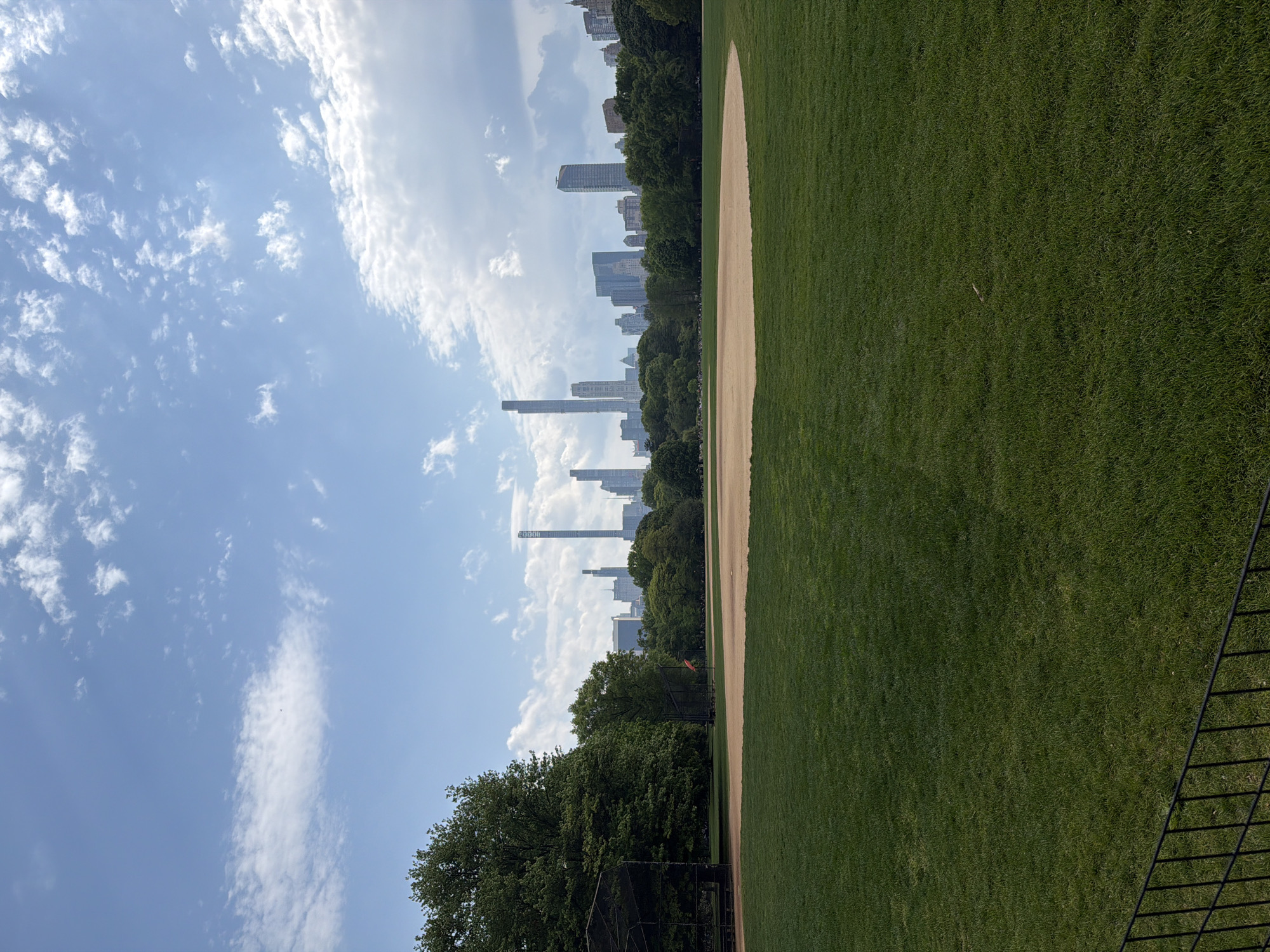 The image shows a baseball field in New York City's Central Park, with a clear blue sky filled with scattered clouds overhead. A line of trees separates the field from the city skyline in the distance, which features several tall buildings. The baseball diamond's infield is composed of tan dirt, contrasting with the green grass surrounding it. A black fence is visible in the bottom right corner of the frame.