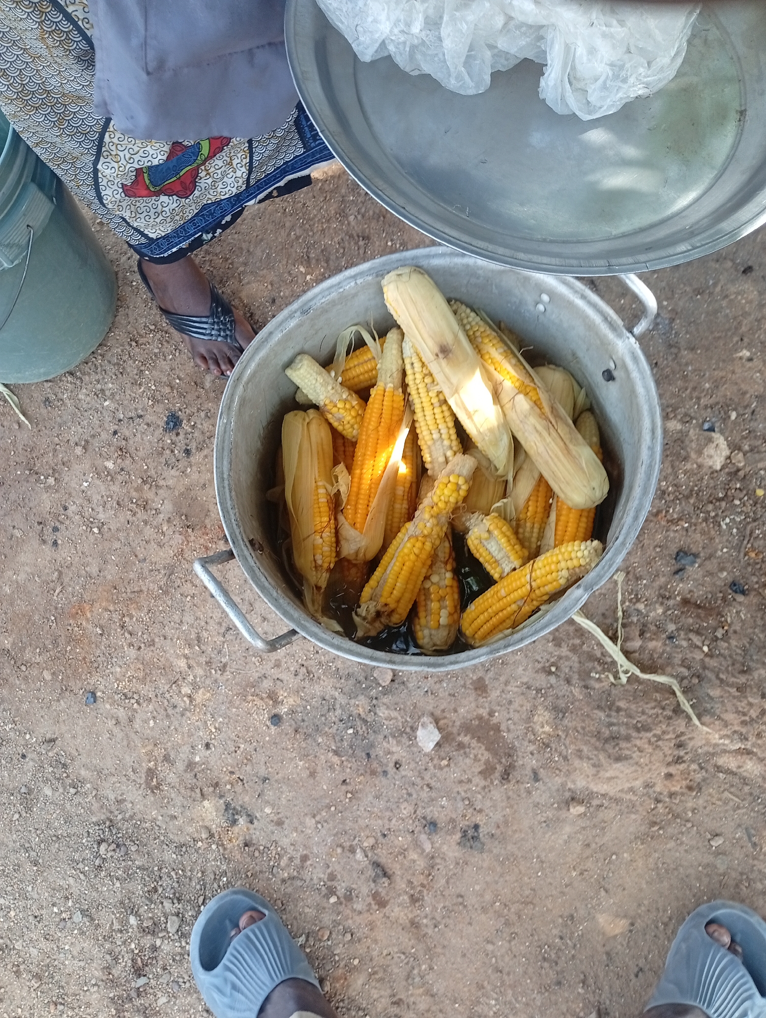 An overhead, eye-level shot shows a metal pot full of corn on the cob, likely being boiled, sitting on a sandy, dirt ground. The pot has two handles and is about one-third full of corn, some of which are submerged in water. The corn cobs are yellow with husks partially removed, exposing the kernels. The ground is a mix of sand and small pebbles, with some sparse, dry vegetation and a few small, dark specks that could be debris or ash. To the left of the pot, a light gray plastic bucket is partially visible. In the background, a person's legs are visible, wearing a patterned skirt and black flip-flops. The top right corner of the image shows a portion of a large, shallow metal dish with a white, crinkled plastic bag inside. The scene is outdoors, possibly in a market or an area where food is being prepared. The location is identified as Rafin Par, Nigeria.