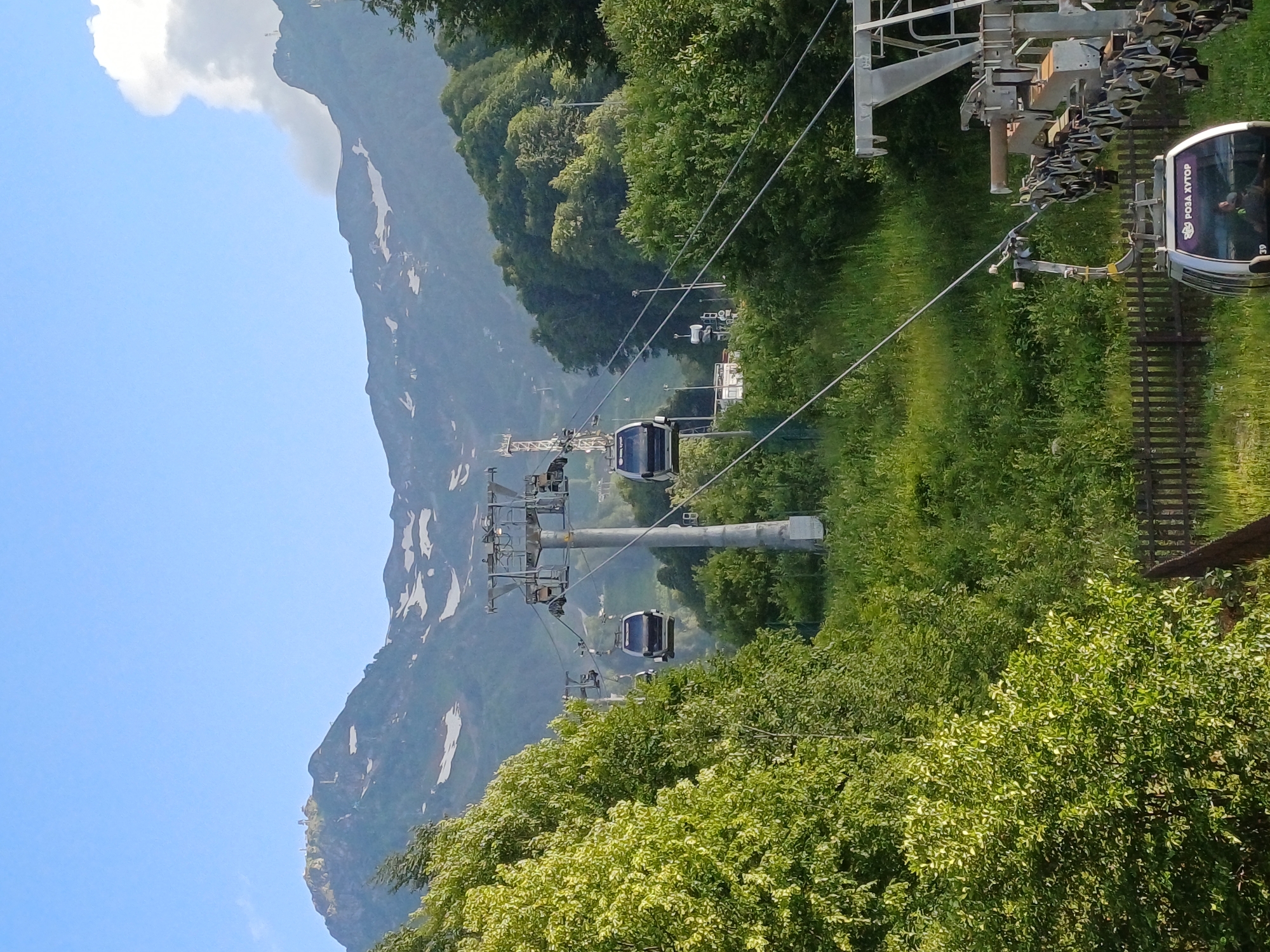 In the mountain resort of Rosa Khutor, Estosadok, Russia, a cable car system transports passengers through a lush, green landscape. Multiple cable cars, some appearing to be in motion, ascend the mountain slopes, their cabins sleek and modern. They are suspended from thick steel cables that stretch between towering pylons. The surrounding environment is characterized by dense vegetation, with vibrant green trees and foliage dominating the foreground and midground. In the background, majestic mountains rise, their peaks partially covered in snow, hinting at the higher altitudes. The sky is a clear, pale blue, with a few wispy clouds near the mountain peaks. The image captures a serene and scenic view of a mountain resort, likely during the warmer months given the verdant landscape.