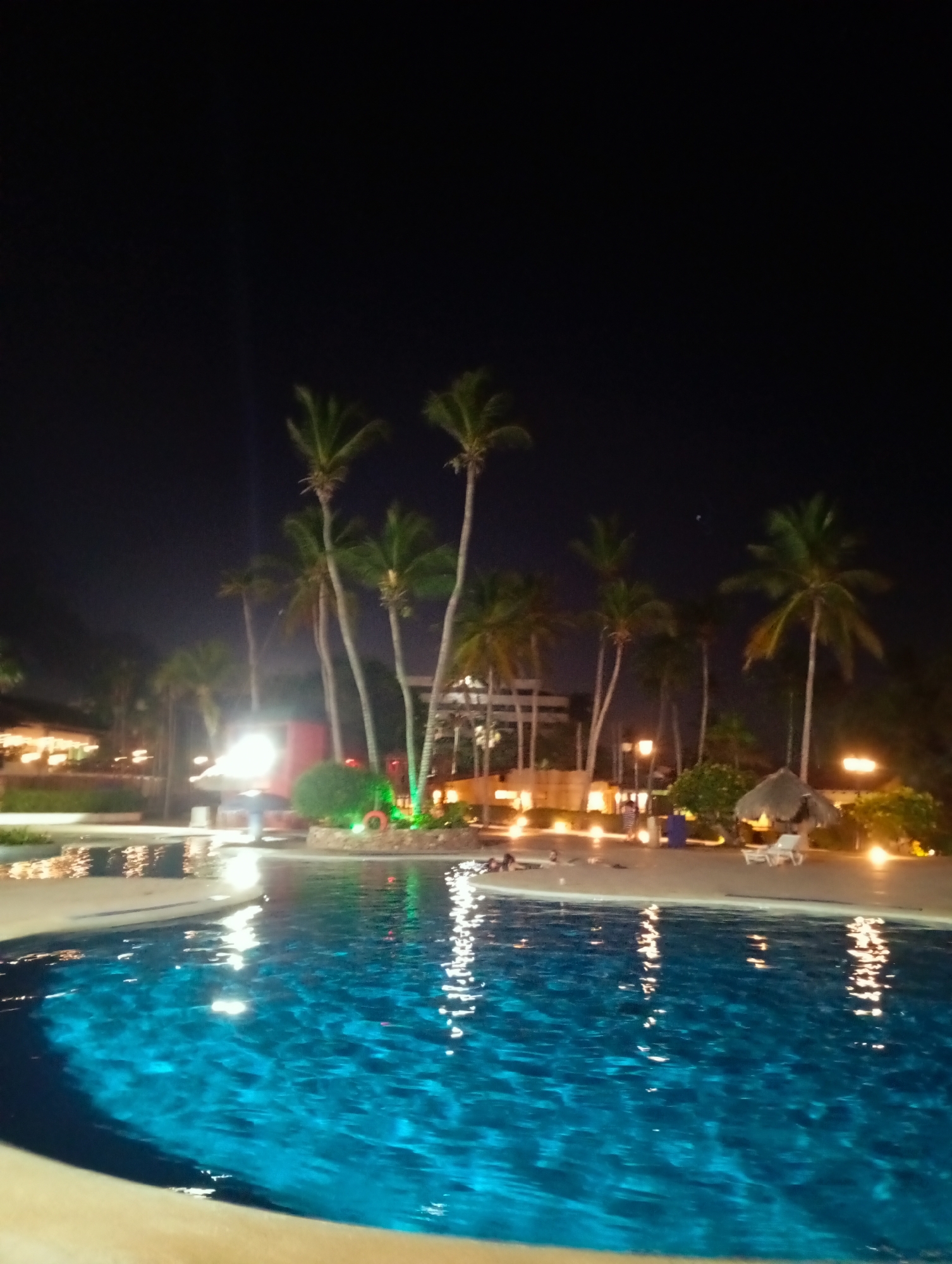 The image shows a night scene at a resort in Playa El Agua, Venezuela. A large swimming pool with turquoise water fills the foreground, reflecting light. Behind the pool, several palm trees line the area near the pool deck. There are several light sources on the deck, including small, warm-toned lights illuminating pathways and buildings. A small tiki hut structure with two white chairs underneath is visible on the pool deck to the right. In the background, buildings can be seen, with lights inside suggesting activity. The sky above is dark and clear. A red life ring is installed on one of the greenery shrubs. A few people can be seen on lounge chairs, relaxing near the pool. The image gives the impression of a calm and relaxing atmosphere.