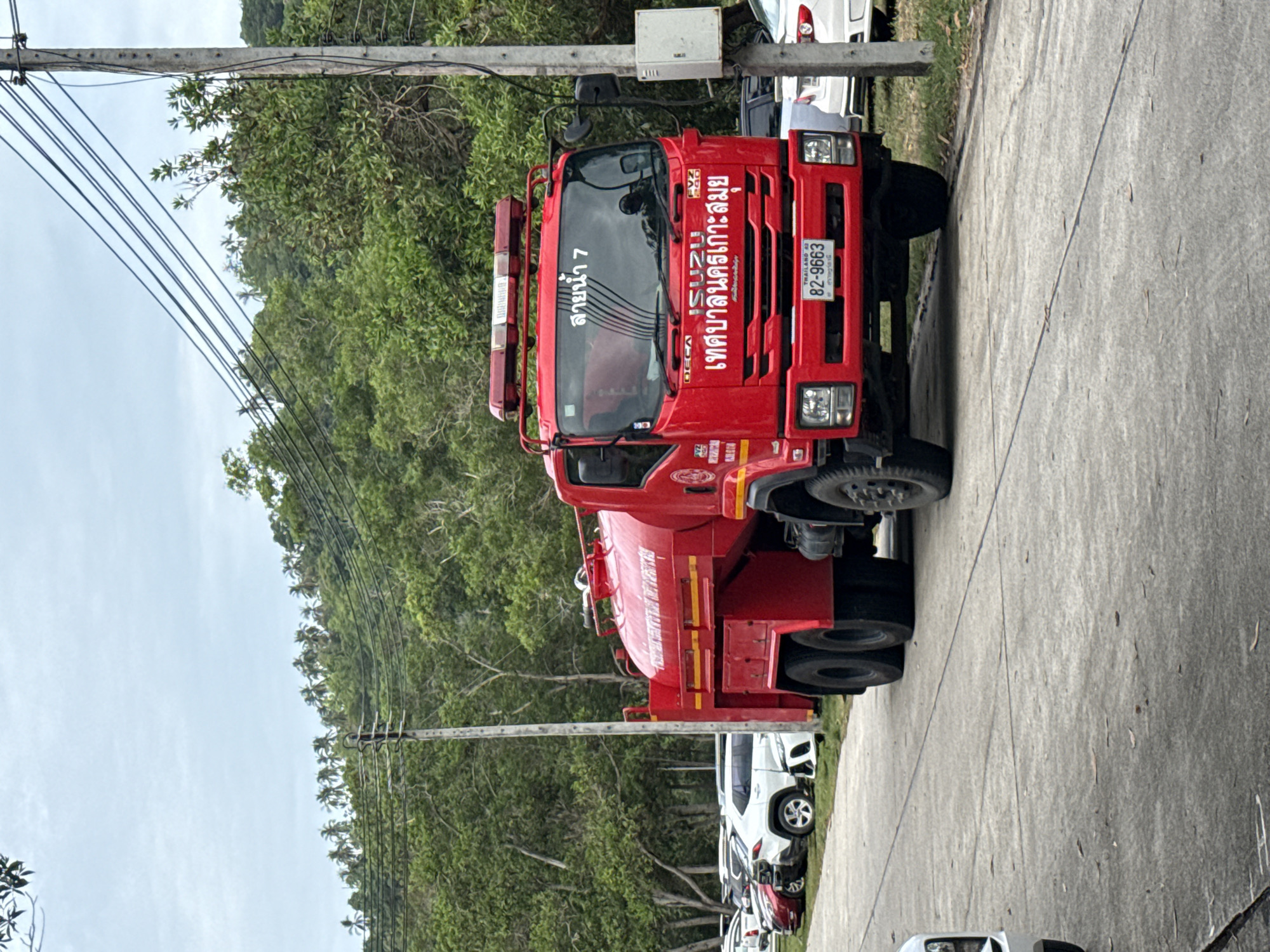 A red fire truck with the words "เทศบาลนครเกาะสมุย" (Koh Samui Municipality) and "สายน้ำ 7" (Water Line 7) written on it is parked on a paved road in a tropical environment. The truck has the license plate "82-9663". Several other vehicles are parked behind the fire truck, and lush green trees and palm trees are visible in the background, with a cloudy sky overhead. A utility pole with wires is to the right of the truck. The scene appears to be an outdoor parking area or roadside in Koh Samui District, Thailand.