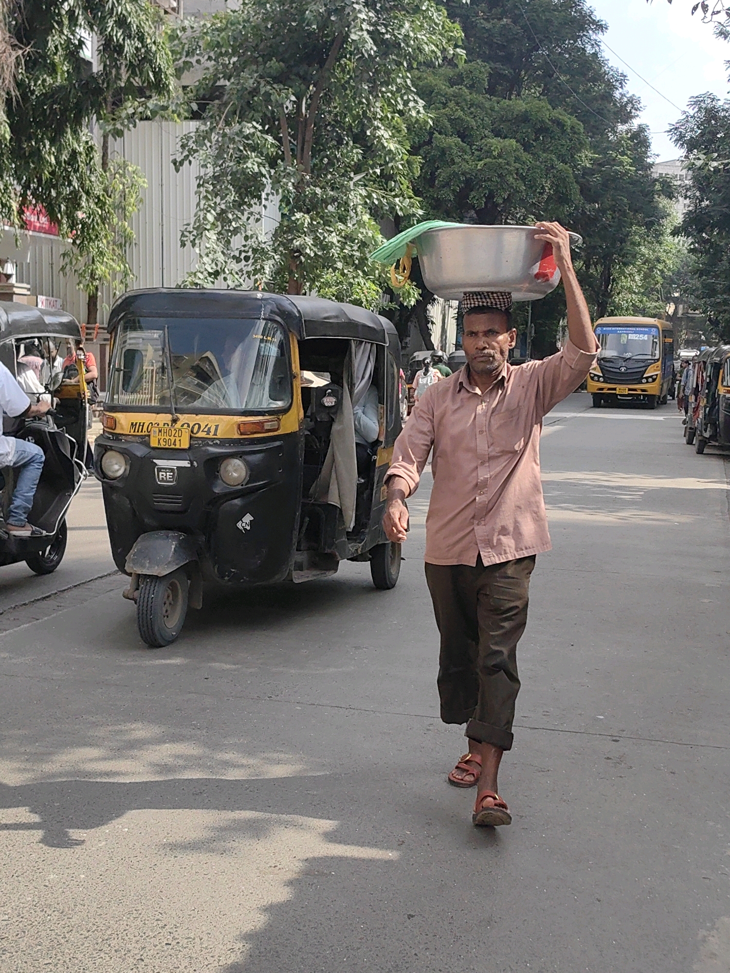 A man is walking on a street in Mumbai, India, carrying a large metal bowl on his head. He is wearing a light brown shirt and dark pants. An auto-rickshaw is parked next to him on the left. In the background, there are trees, buildings, and a yellow bus. The scene appears to be during the daytime, with sunlight casting shadows on the road.