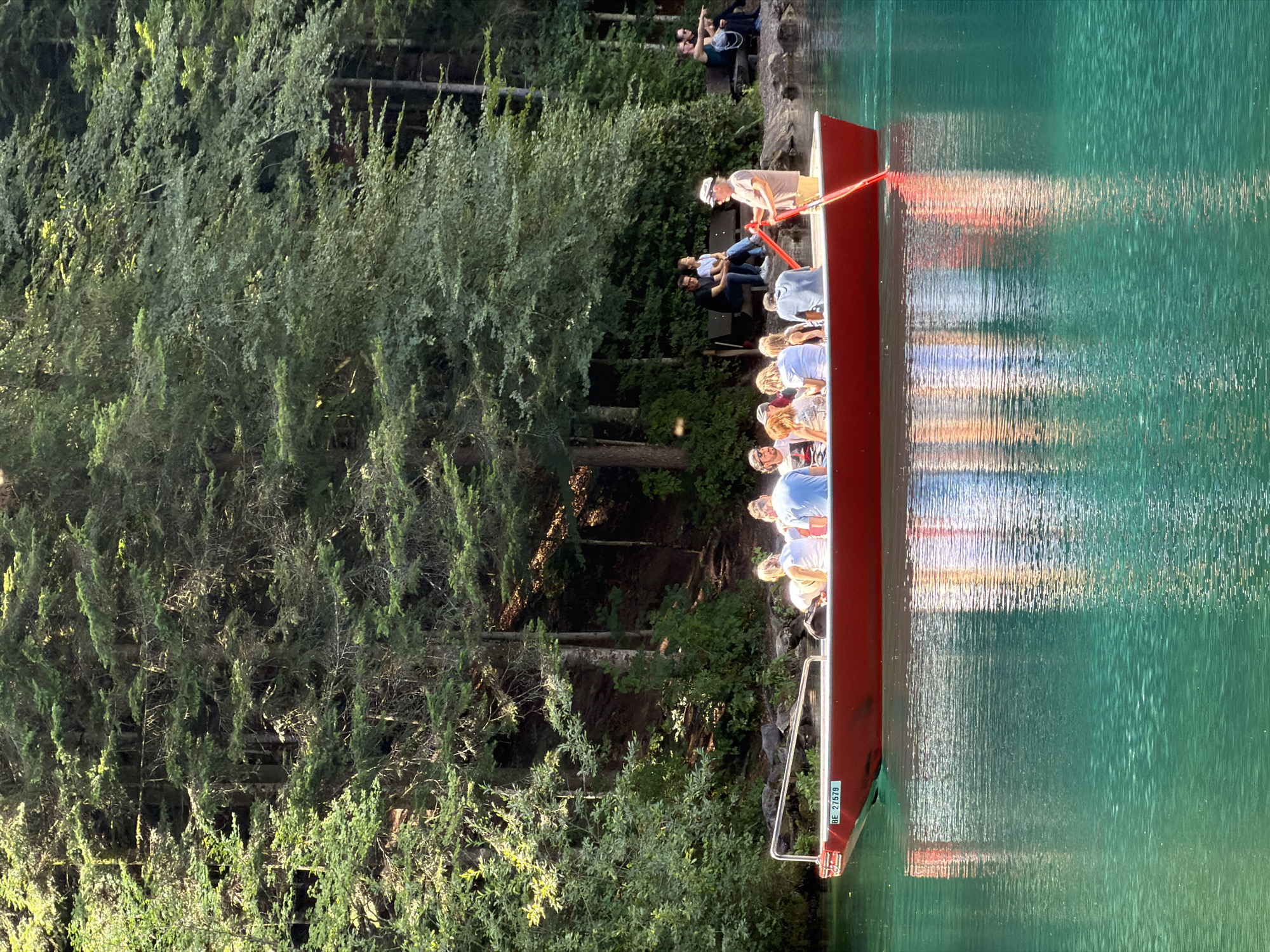 A red boat with a license plate reading "BE 27579" travels across a vibrant turquoise lake. The boat is filled with passengers, most of whom are facing forward. One individual, wearing a hat and holding an oar, is rowing the boat. The background features a dense forest with tall green trees and dappled sunlight. To the right of the lake, several people are seated on benches, observing the scene. The overall atmosphere suggests a leisurely outing in a natural setting, likely the Blausee Lake in Kandergrund, Switzerland, known for its clear, colorful water and surrounding forests.