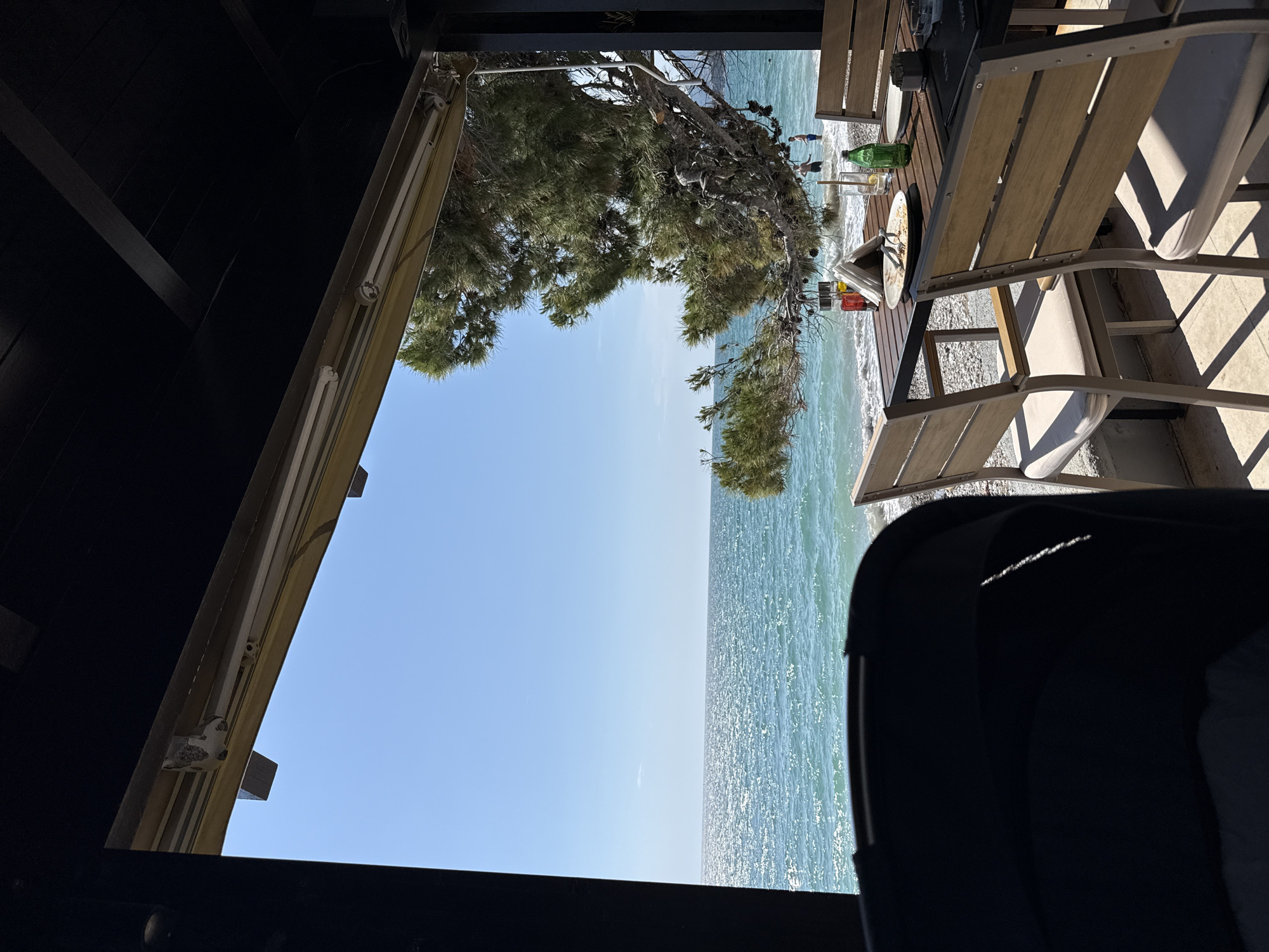 View from under a dark structure toward the sea. Clear blue sky and aquamarine water visible. A tree branch hangs partially in view. On a wooden deck, a dining table is set with a bottle, plates, napkins, and cutlery. Beach chairs sit around the table. Figures of people can be seen on the beach in the distance. The scene is located in the city of Bar, Montenegro.