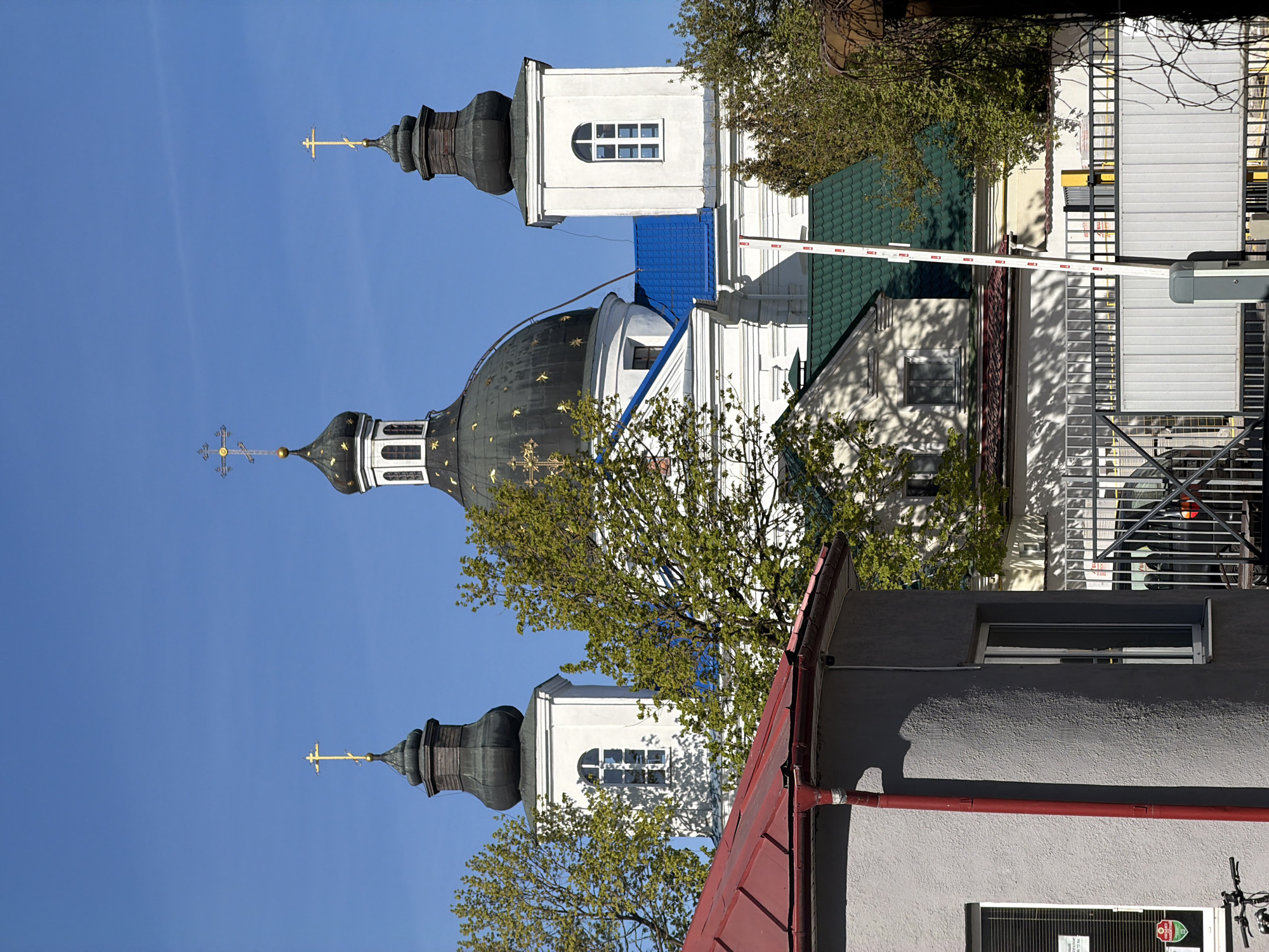 The image shows a low-angle view of a church with three towers, featuring a dome and Orthodox crosses, located in Hrodna, Belarus. The church is white with dark roofs. It is positioned against a bright blue sky. Parts of adjacent buildings are visible, including a gray building with a red roof and a white building with a green roof. A boom barrier is also visible. Trees with green leaves partially obscure the view of the buildings.