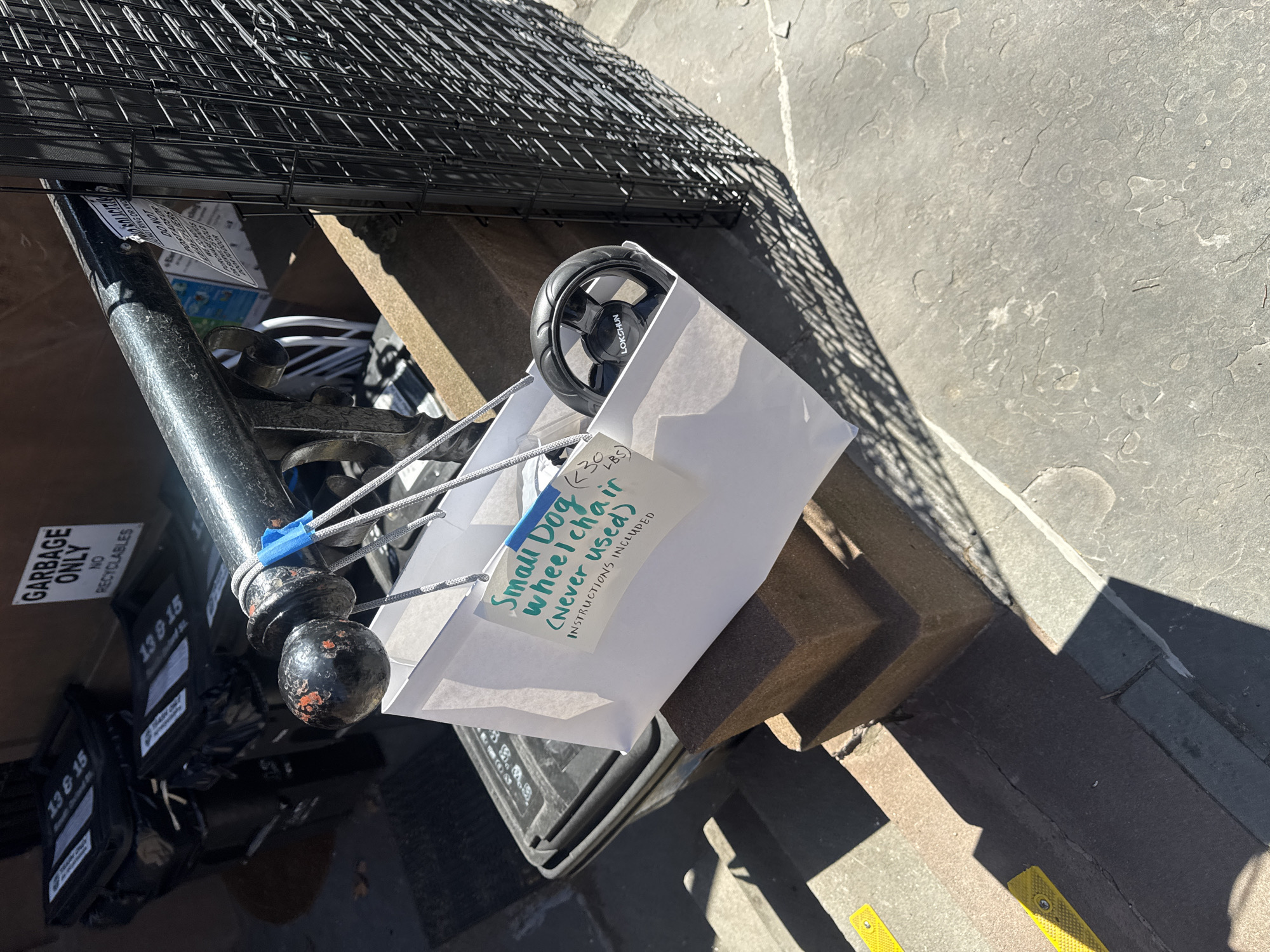 The image shows a discarded "Small Dog Wheelchair" in a white paper bag, with "Never used" and "Instructions included" written on a label affixed to the bag. The bag is hanging from a black metal post with a rounded top, which is part of a fence or barrier alongside a trash receptacle. A black wheel protrudes from the bag, indicating that the wheelchair is inside. The trash receptacle has a "Garbage Only, No Recyclables" label and is marked with "13 & 15" and "13&15" on the bins. The background consists of a concrete surface, possibly a sidewalk or the edge of a building. The scene appears to be outdoors, with a shadow cast on the ground. No people are visible.
Given the "13 & 15" markings on the trash cans, which denote specific sanitation districts in New York City, along with the style of the trash receptacle and the urban setting, the location is likely somewhere within the city of New York, United States of America.