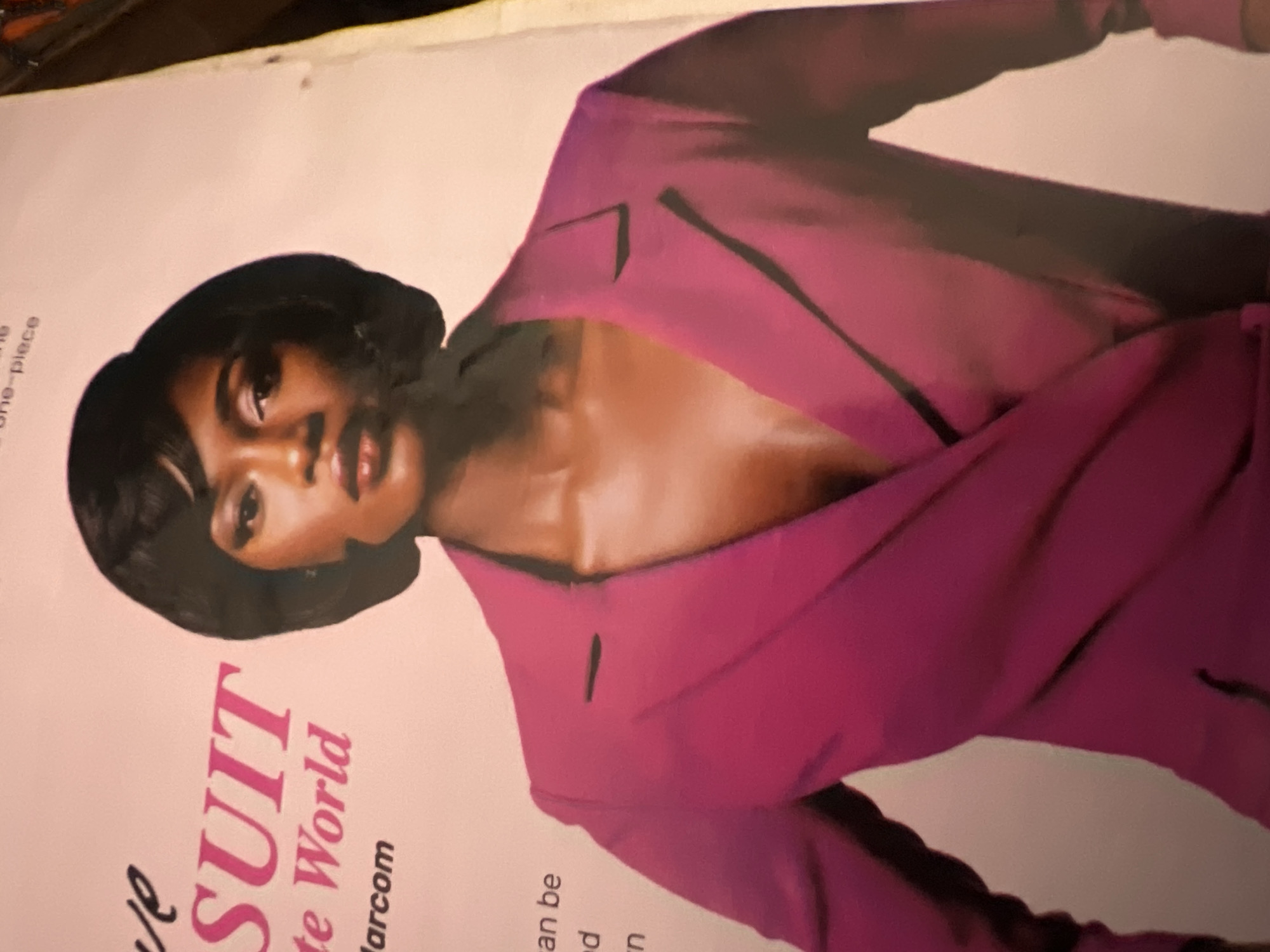 The image is a close-up portrait of a woman wearing a magenta blazer. The woman has dark skin, dark hair styled in a bob, and is looking directly at the camera with a serious expression. The blazer is double-breasted with wide lapels, and the background is a pale pink. Text is visible on the left side of the image, with words like "SUIT," "World," and "larcom" appearing to be part of a magazine or advertisement. The image appears to be a photograph of a printed page, possibly a magazine spread, as indicated by the texture and lighting. There is no discernible information or imagery within the photograph that relates to the city of Jalingo, Nigeria.