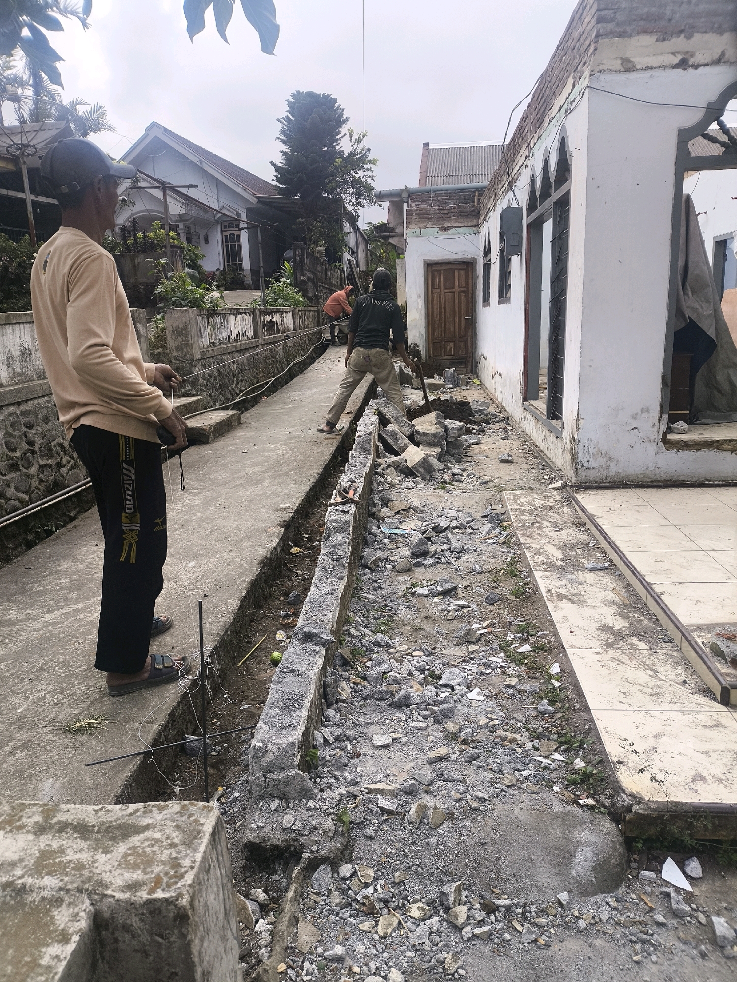 The image depicts a street scene in Kandangtepus, Indonesia, where construction or demolition is underway. In the foreground, a person wearing a beige long-sleeved shirt and black pants with yellow stripes stands facing left, holding a string and some tools. To the right and further down the street, another person in a black shirt and khaki pants is using a shovel to break up debris. A damaged building with exposed brickwork and missing walls is visible on the right side of the image. The ground is covered with rubble and dirt. The environment appears to be a residential area with houses visible in the background. The sky is overcast.