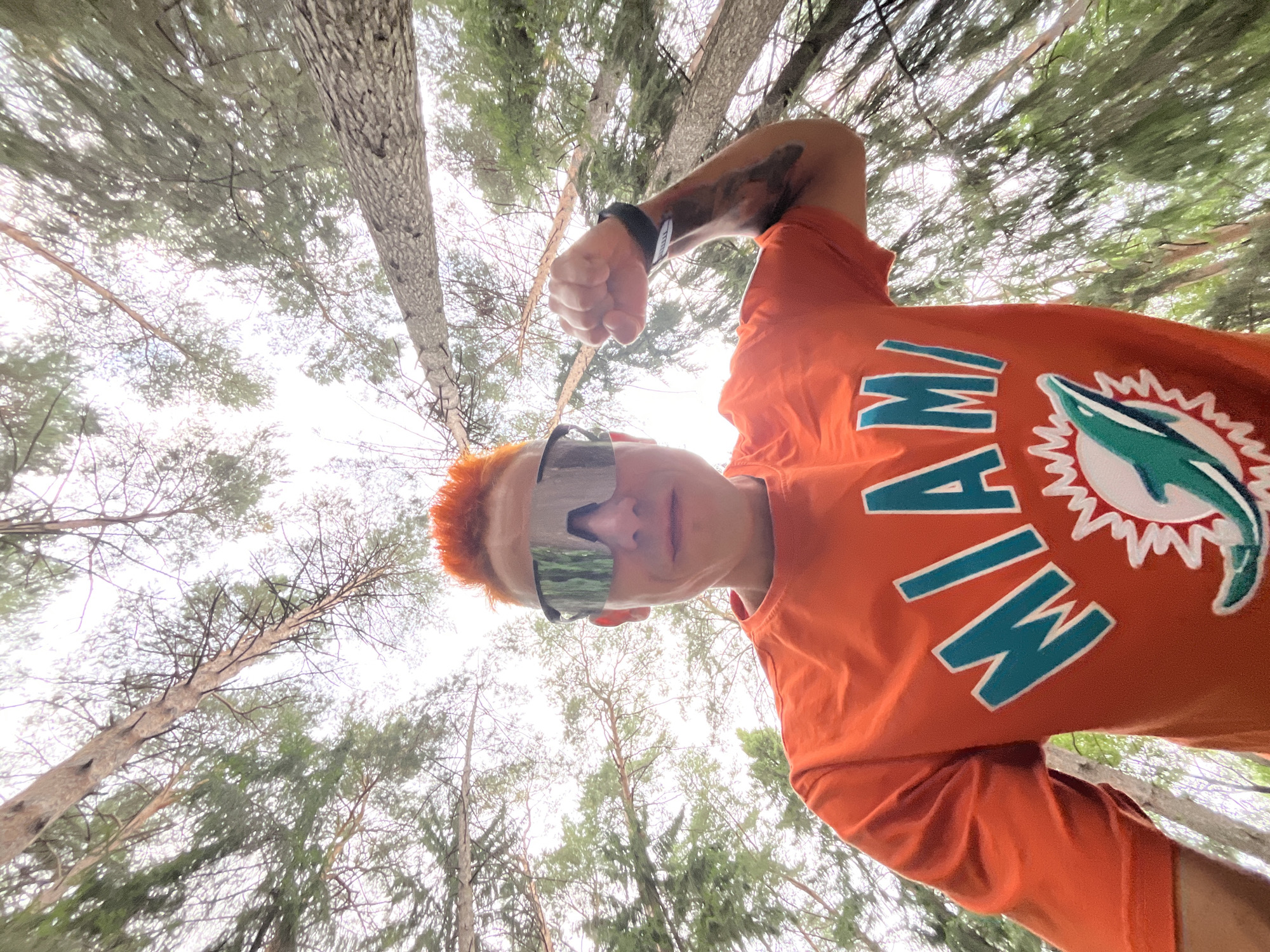 A low-angle, wide shot shows a person with bright orange hair, wearing reflective sunglasses and an orange t-shirt with the word "MIAMI" in teal lettering and a dolphin logo, flexing their bicep. They are outdoors in a forest, looking up at the camera. Tall, thin trees with green foliage fill the background, and the sky is overcast and bright. The location of this image is the city of Chaikovsky, Russia.