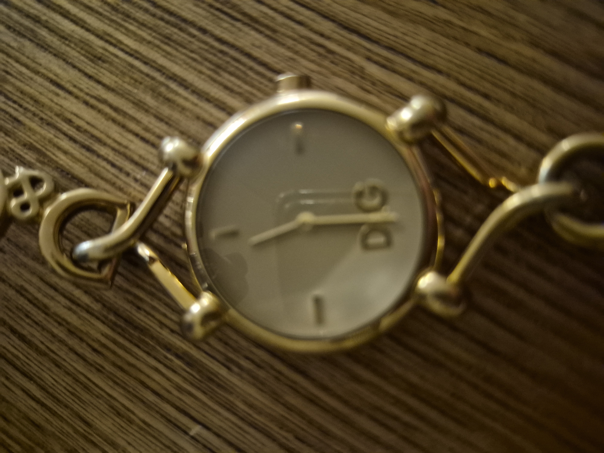 A gold Dolce & Gabbana watch with a chain band is resting on a wooden surface. The watch face is circular and off-white with gold hour markers. The Dolce & Gabbana logo is visible on the watch face. The watch hands are gold and appear to be pointing to approximately 1:15. The background is a close-up, slightly out-of-focus shot of wood grain.