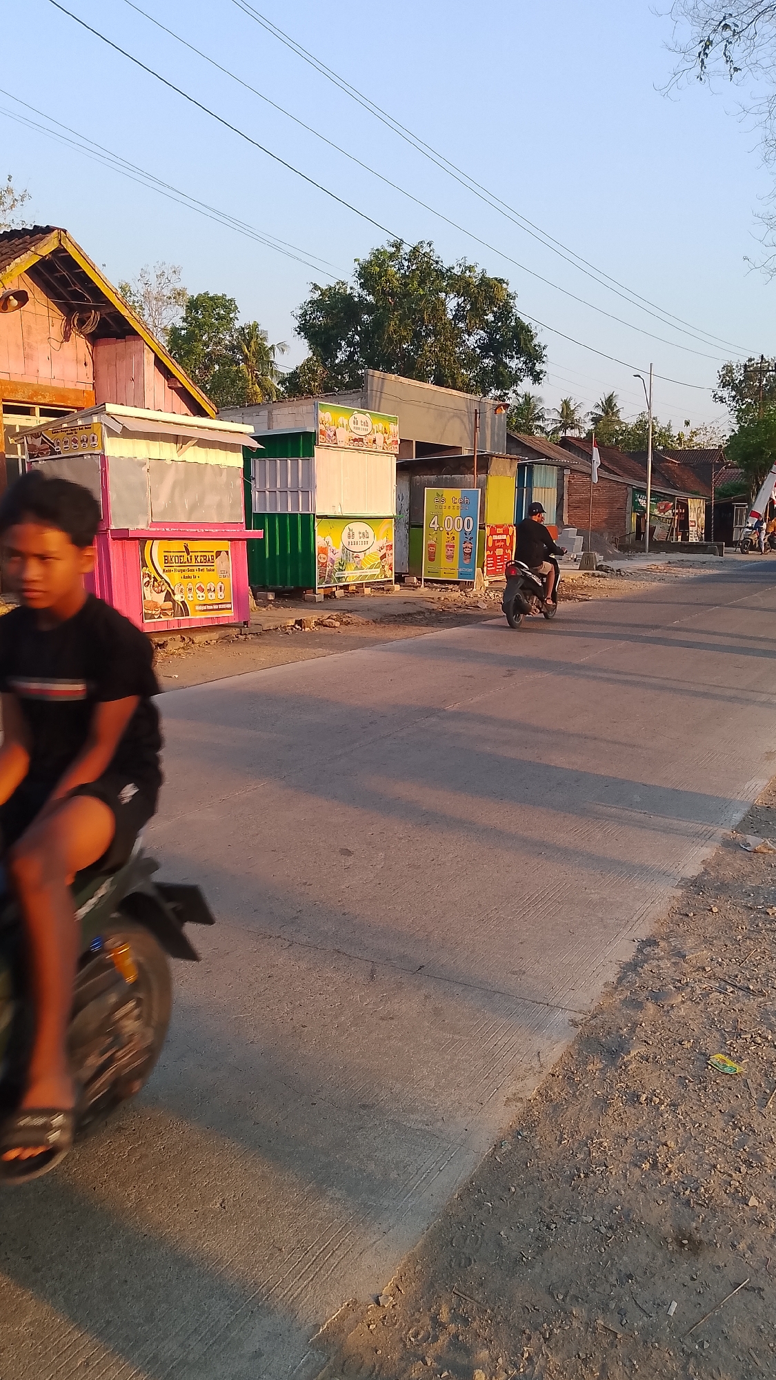 This image depicts a street scene in Karanglangu, Indonesia, characterized by informal roadside stalls and a dusty, sunlit environment. In the foreground, a young man is riding a motorcycle, his body slightly blurred by motion. The motorcycle is dark-colored, with a prominent rear wheel and fender. The road surface is concrete, showing tire marks and patches of dirt.
The background features several brightly colored food stalls, likely serving local cuisine. One stall is painted pink and displays a sign with text including "Bikoli Kebab". Another stall is green, and further down the street, a yellow stall has a sign advertising "Es Teh" (iced tea) for 4,000 Indonesian Rupiah, with images of beverages. Beyond the stalls, low-rise buildings and trees are visible, under a clear blue sky. Power lines are strung across the upper portion of the image. Another person is riding a motorcycle further down the street, heading away from the camera, and several other figures are present near the stalls. The overall impression is one of a bustling, everyday scene in a semi-urban or rural Indonesian setting during daylight hours, with warm, long shadows suggesting late afternoon or early morning sun.