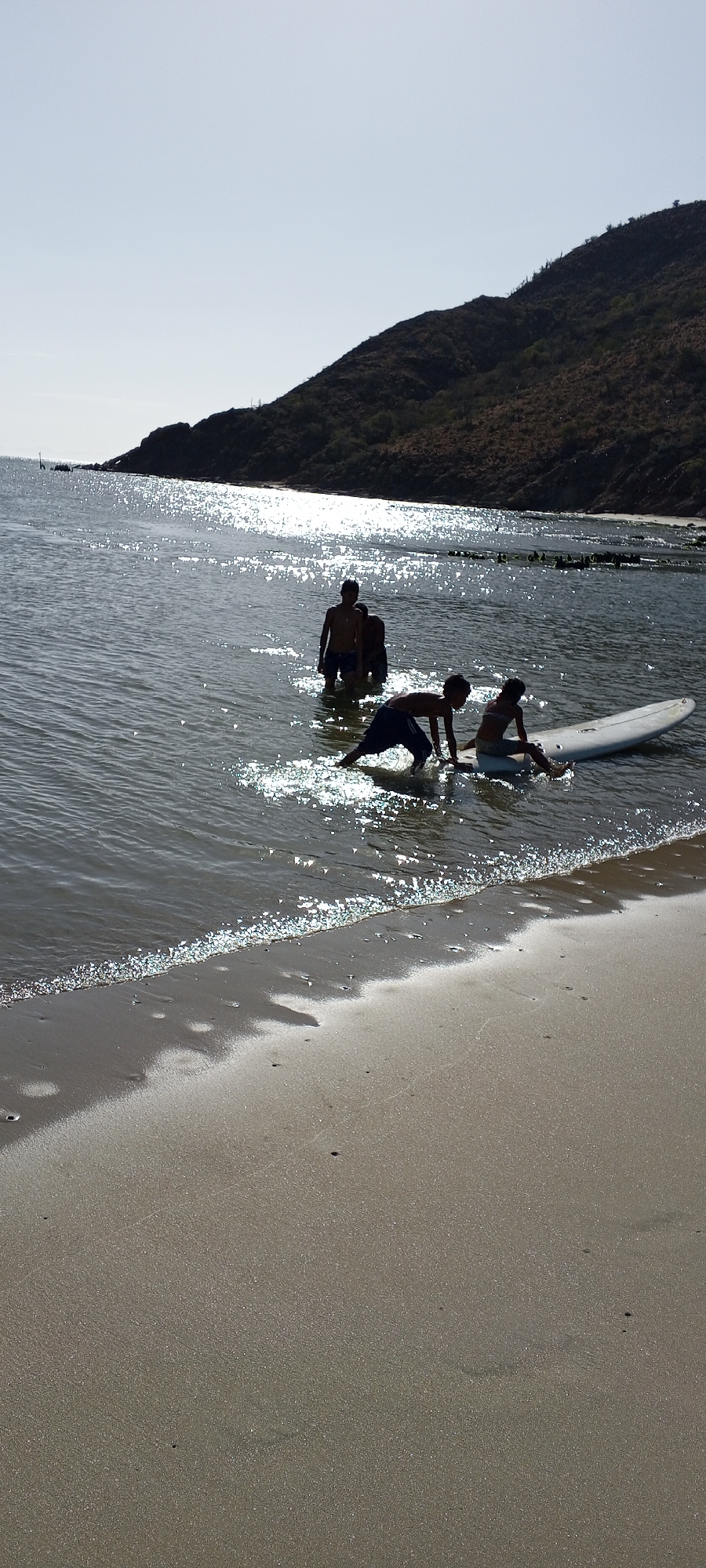 The image depicts a beach scene. In the water, three figures are standing and another two figures are interacting with a surfboard. The background includes a hill/mountain. The location is Unknown, Unknown.