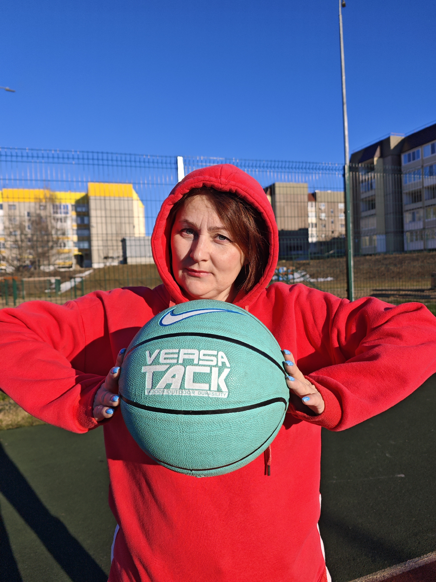 A woman in a red hooded sweatshirt is holding a teal basketball with "VERSA TACK" written on it. She is standing on a dark green surface that appears to be an outdoor sports court. A fence is visible behind her, along with buildings that suggest a residential area. Based on the location identified as Markovskii, Russia, the buildings could be apartment blocks. The sky is clear and blue, indicating a sunny day. The woman's fingernails are painted in a similar teal shade to the basketball. A streetlight is visible in the background. The overall scene appears to be a casual portrait taken on a basketball court in a residential area.