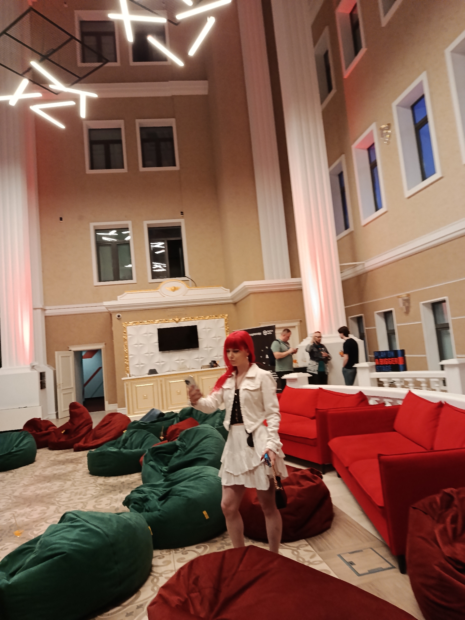 The image captures an interior setting. The space is furnished with red sofas and an assortment of green and red beanbag chairs, arranged on a patterned carpet. A woman with red hair stands amidst the seating arrangement, holding a phone or device in her hand, appearing to take a photo or video. In the background, three individuals are conversing near a white railing. A large, white decorative frame with a television screen is situated along the back wall. The interior architecture includes high ceilings, tall pillars, and windows, suggesting a spacious, possibly public venue in the city of Unknown, Unknown. The lighting fixtures are modern and contribute to the ambiance.