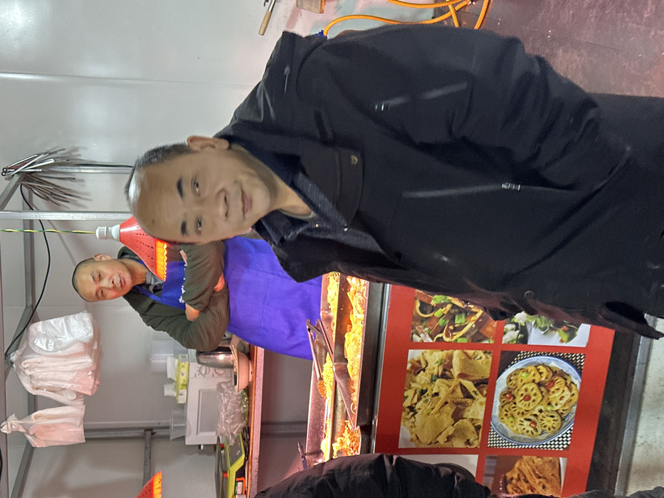 An indoor scene in Jiangjin, China, depicts a food stall or eatery. In the foreground, an East Asian male, approximately middle-aged, is positioned slightly to the right of the frame, facing the camera. He wears a dark, possibly black, padded jacket over a patterned collared shirt. His hands are out of view, possibly in pockets. Behind him, another East Asian male, also middle-aged with a shaved head, stands behind a stainless steel counter. He is wearing a dark green jacket over a striped shirt and a blue apron, with his arms crossed over his chest. He looks towards the camera.
The counter behind the foreground subject holds several rectangular stainless steel trays containing various cooked food items, illuminated by two red, cone-shaped heat lamps suspended from above by green and yellow wires. Above the counter, a microwave oven, an electric kettle, a food scale, and various white bowls and containers are visible on a lower shelf. White plastic bags or gloves are hanging from a metal rack at the top left. Another metal rack to the right of the background man holds several metallic utensils. The lower portion of the food stall features display panels with images of prepared dishes, including one with flatbreads, another with stir-fried items, and a prominent image of sliced lotus root. A yellow hose or cable is visible on the floor to the far right. The walls of the stall appear to be white or light-colored, possibly made of metal panels, and the floor is a dark color. A person's arm, dressed in dark clothing, is partially visible on the far left edge of the frame.