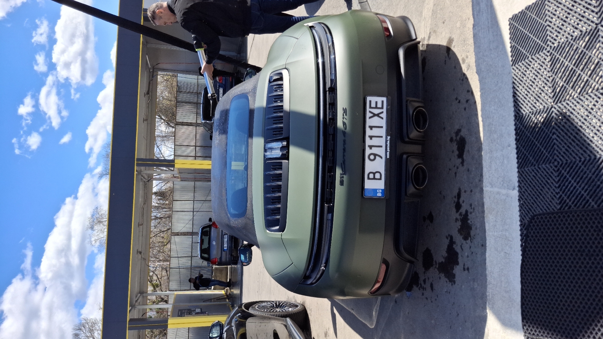 The image depicts the rear of a matte olive green Porsche 911 Carrera 4 GTS parked at a car wash in Varna, Bulgaria. The license plate reads "B 9111 XE." A person is washing the car with a pressure washer. The car is parked on a wet surface next to a textured, non-slip mat. Another car is visible in the background, with a license plate that begins with "B 327." The sky is partly cloudy and blue. The car wash has a metallic frame and roof.