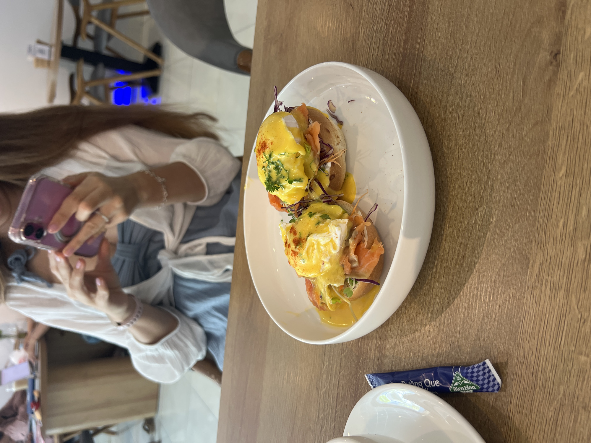 The image shows a plate of Eggs Benedict with salmon on a wooden table. A woman wearing a blue dress and a white cardigan is taking a photo of the food with her phone. A sugar packet labeled "Bien Hoa" is visible next to the plate. The location is likely a restaurant in Bắc Nha Trang, Vietnam.