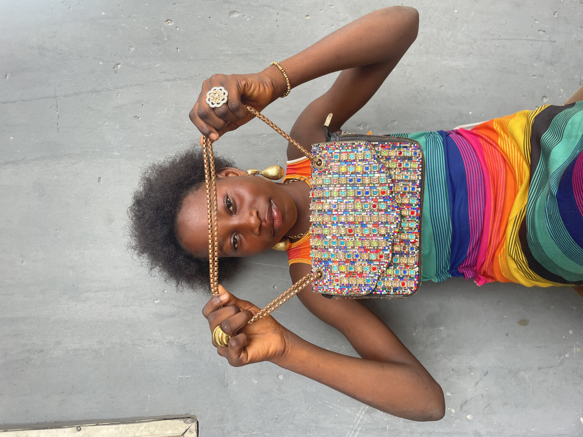 A young person with dark skin and dark, curly hair is depicted lying on their back on a textured grey surface, which appears to be concrete ground. Their head is tilted, and they are looking directly at the camera with a neutral and engaging expression.
The individual is wearing a vibrant, multi-colored striped garment, likely a dress or top, showcasing broad vertical stripes in shades of red, orange, yellow, green, teal, and blue. They are adorned with several pieces of gold-colored jewelry: a prominent floral-shaped ring with sparkling details on their right ring finger, a simpler stacked ring on their left ring finger, a beaded bracelet on their right wrist, and large teardrop-shaped earrings.
With both hands, the person is holding the gold-colored chain strap of a rectangular handbag, presenting it towards the viewer. The handbag itself is a key focal point, richly decorated with numerous small, brightly colored jewels or sequins arranged in a grid pattern, creating a dazzling, multi-hued effect across its surface.
The lighting is bright and even, indicating daytime conditions. The background is an unadorned grey surface, offering no specific architectural or natural features. The setting is simple, likely an outdoor or utilitarian indoor space, consistent with a casual portrait. This scene takes place in Badagry, Nigeria.