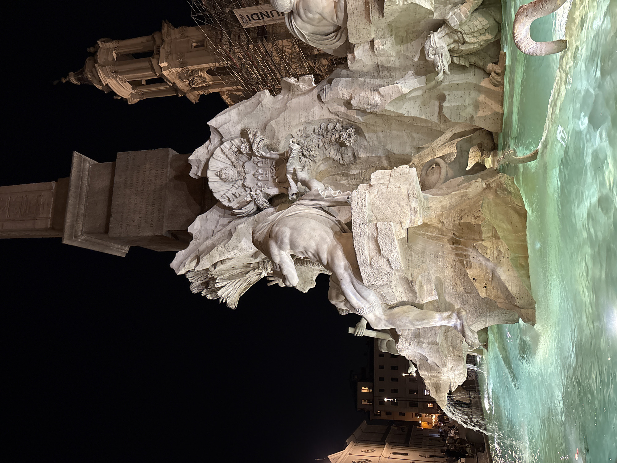 This is a nighttime photograph of the Fountain of Four Rivers in Piazza Navona, Rome, Italy. The central subject is the monumental Baroque fountain, which features a large sculpture of a muscular male figure representing the Nile River, holding an oar and his arm raised. Surrounding him are other sculpted figures and animals, including a crocodile and a lion, all carved from white marble. The fountain's basin is filled with illuminated, turquoise water, with cascades of water flowing over the sculpted rocks. Behind the fountain, a tall, ancient obelisk rises into the dark night sky. To the right of the obelisk, a church tower with a dome and ornate architectural details is visible, with scaffolding attached to its side, suggesting ongoing construction or restoration work. In the background, beyond the fountain, the facades of buildings with lit windows are dimly visible. Several people are present in the scene, standing or walking in front of the fountain and along the edge of the piazza. The overall atmosphere is dramatic, illuminated by artificial lighting that highlights the sculptures and the water.