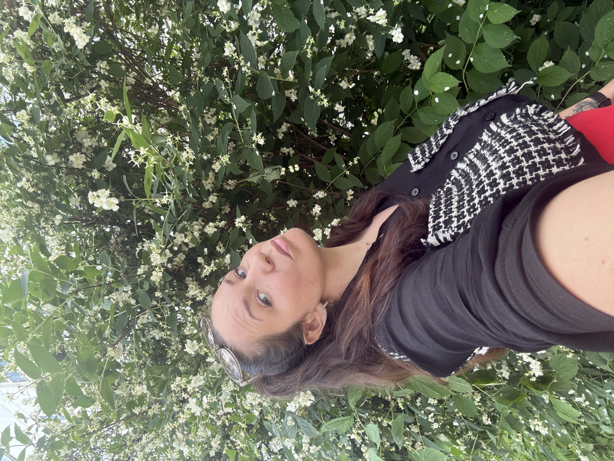 In Chaikovsky, Russia, a woman is taking a selfie in front of a flowering bush. She is wearing a black shirt with a houndstooth vest over it and red pants. Her brown hair is pulled back in a ponytail, and she has sunglasses on top of her head. The bush is covered in small white flowers and green leaves, suggesting it is springtime or early summer. The lighting indicates it is daytime, likely outdoors.