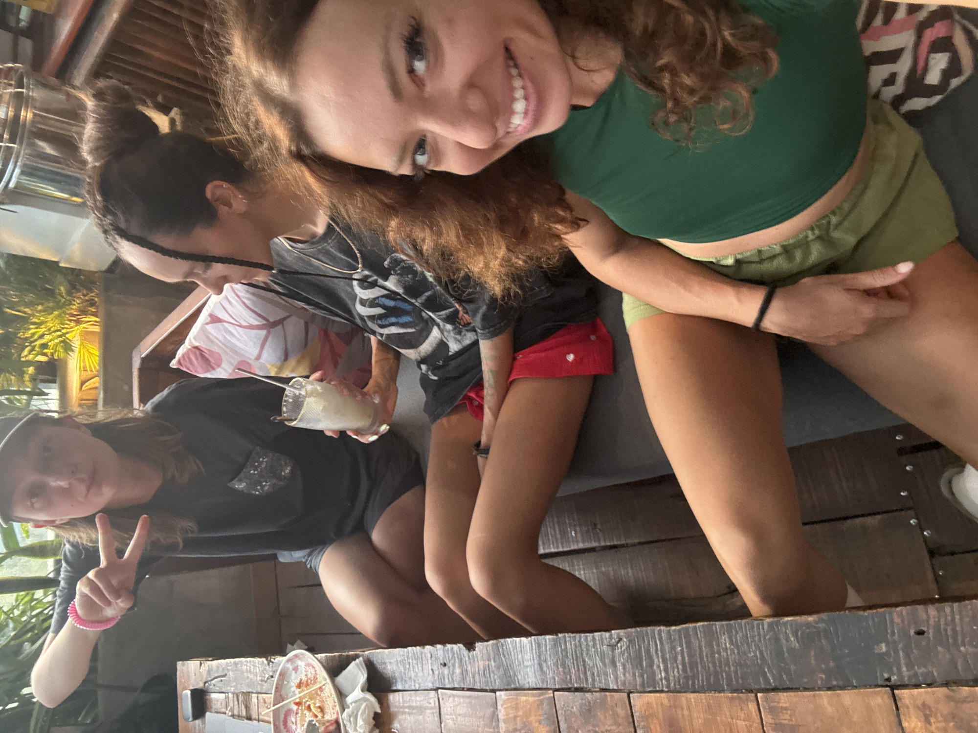 Three females are posing for a selfie inside a building in Hà Giang 1 Ward, Vietnam. The female to the right has curly, shoulder-length brown hair, a green top, and green shorts. She has a bracelet on her left wrist and is sitting next to the second female who has her hair up in a bun and is wearing a grey T-shirt and red shorts. The third female is on the left, wearing a dark T-shirt, dark shorts, and a baseball cap. She is holding a glass and is making a peace sign. There is a half-eaten plate on the table in front of them. The background includes a window with vegetation outside and a patterned sofa.