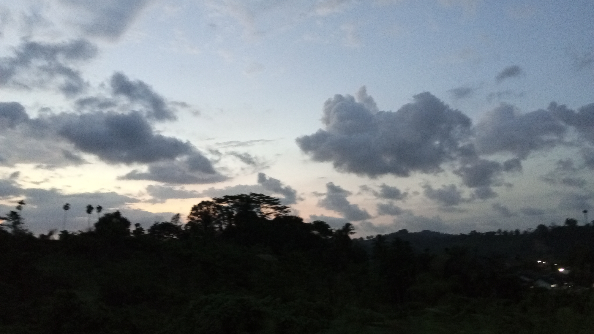 The image shows a landscape in Lhokseumawe, Indonesia, under a cloudy sky at dusk. The foreground consists of dark, silhouetted trees and vegetation covering a hilly terrain. Further in the distance, more hills are visible, also silhouetted against the sky. The sky is filled with varying shades of grey clouds, with a hint of orange light visible near the horizon. In the far distance, a few lights suggest the presence of a town or settlement. There are no people visible in the image.