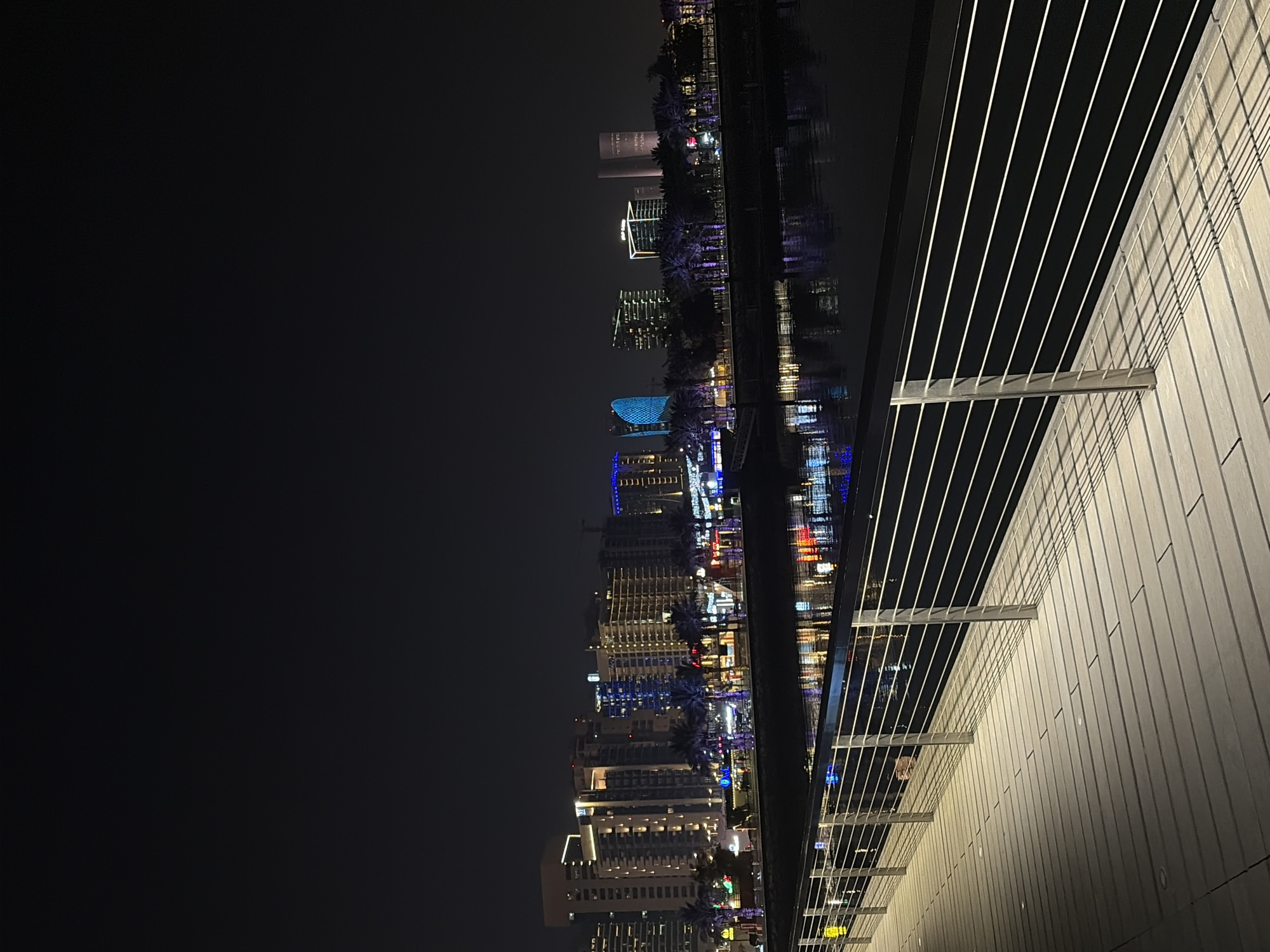 The image shows a nighttime cityscape with a modern boardwalk in the foreground. The boardwalk is made of light-colored wooden planks and is lined with a sleek metal railing. The railing casts long, linear shadows across the boardwalk, indicating the presence of artificial lighting. Beyond the boardwalk, a body of water reflects the lights of the city skyline. The skyline features several tall buildings, some of which are illuminated with colorful lights, including blue and white. Palm trees are visible along the waterfront, adding a tropical element to the urban scene. The overall impression is one of a vibrant and modern waterfront district at night.