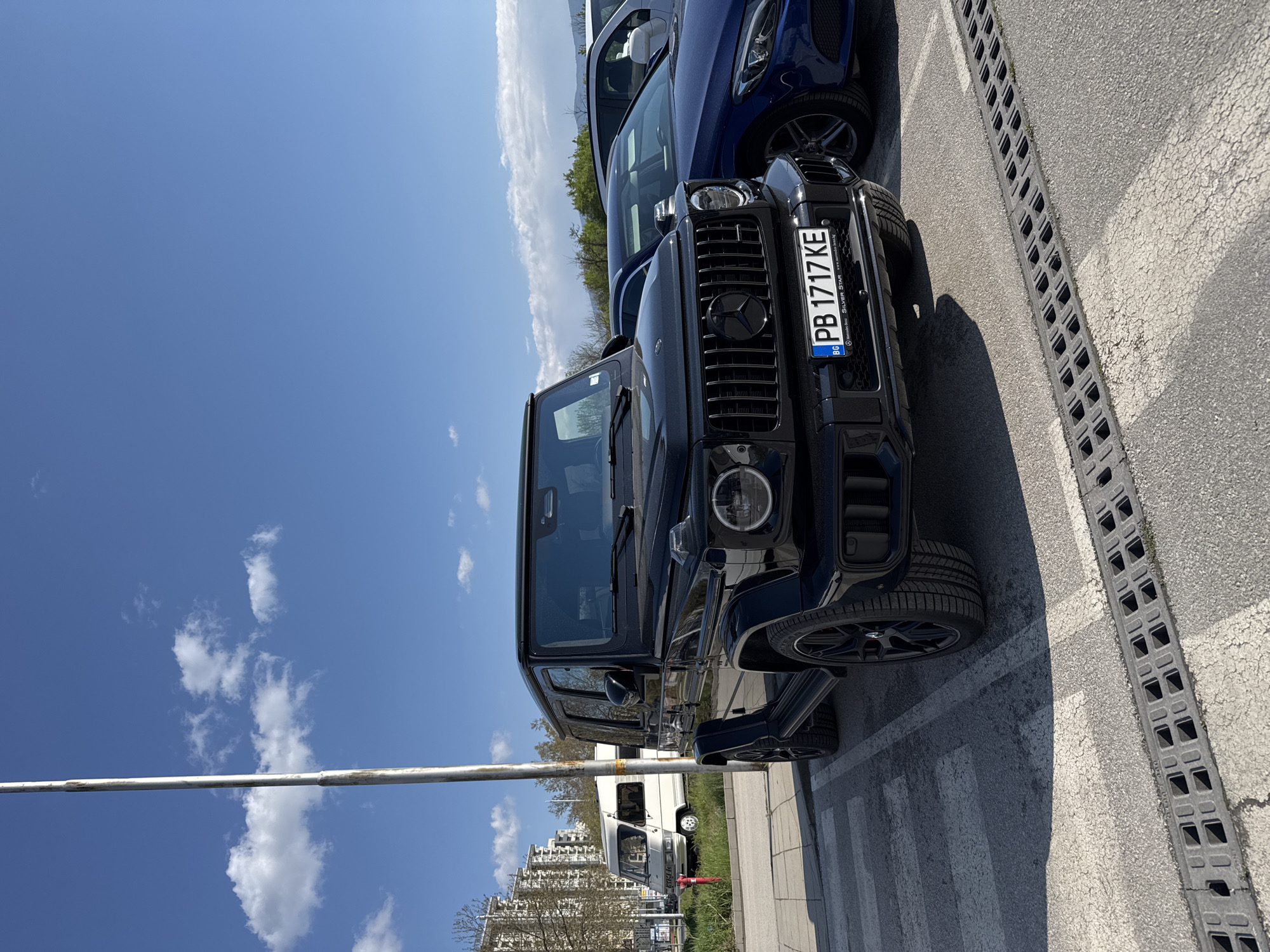 A black Mercedes-Benz G-Class AMG is parked in a designated parking spot. The vehicle has a Bulgarian license plate that reads "PB 1717 KE". Next to the Mercedes-Benz is a dark blue vehicle, partially visible. The parking spot is marked with white lines and has a grey concrete drain running along the side. In the background, there is a white van, some buildings, and trees. The sky is blue with white clouds. The location is in Plovdiv, Bulgaria. The lighting suggests it is daytime.
