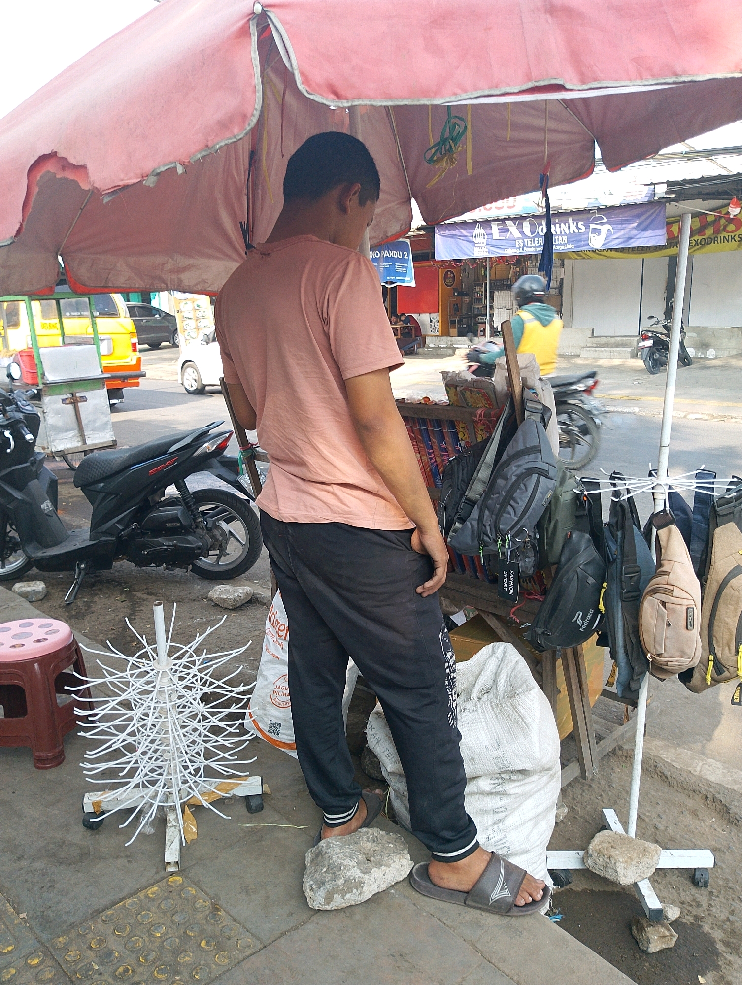 The image captures a street scene in Lembang, Indonesia, under a large, pink umbrella. In the foreground, a person, likely a vendor, stands with their back to the camera, wearing a light pink t-shirt and black pants. They are standing on two rocks, one of which is supporting their right foot. To the left, a small, maroon stool is visible. In front of the vendor is a white rack with numerous thin, white wire arms, resembling a display stand.
Behind the vendor, a wooden cart is laden with various bags, primarily dark-colored fanny packs and crossbody bags, hanging from a rack. A large, white sack is placed on the ground next to the cart.
In the mid-ground, several vehicles are present. A black scooter is parked to the left, and a yellow vehicle, possibly a modified truck or food cart, is further back. A white car is also visible on the street. The background shows a row of shops with signs, including one that reads "EXODRINKS" in blue and white. People can be seen in the background, including a person wearing a yellow vest and helmet, possibly another vendor or a passerby, and other figures inside a shop. The scene appears to be a busy outdoor market or roadside stall area.